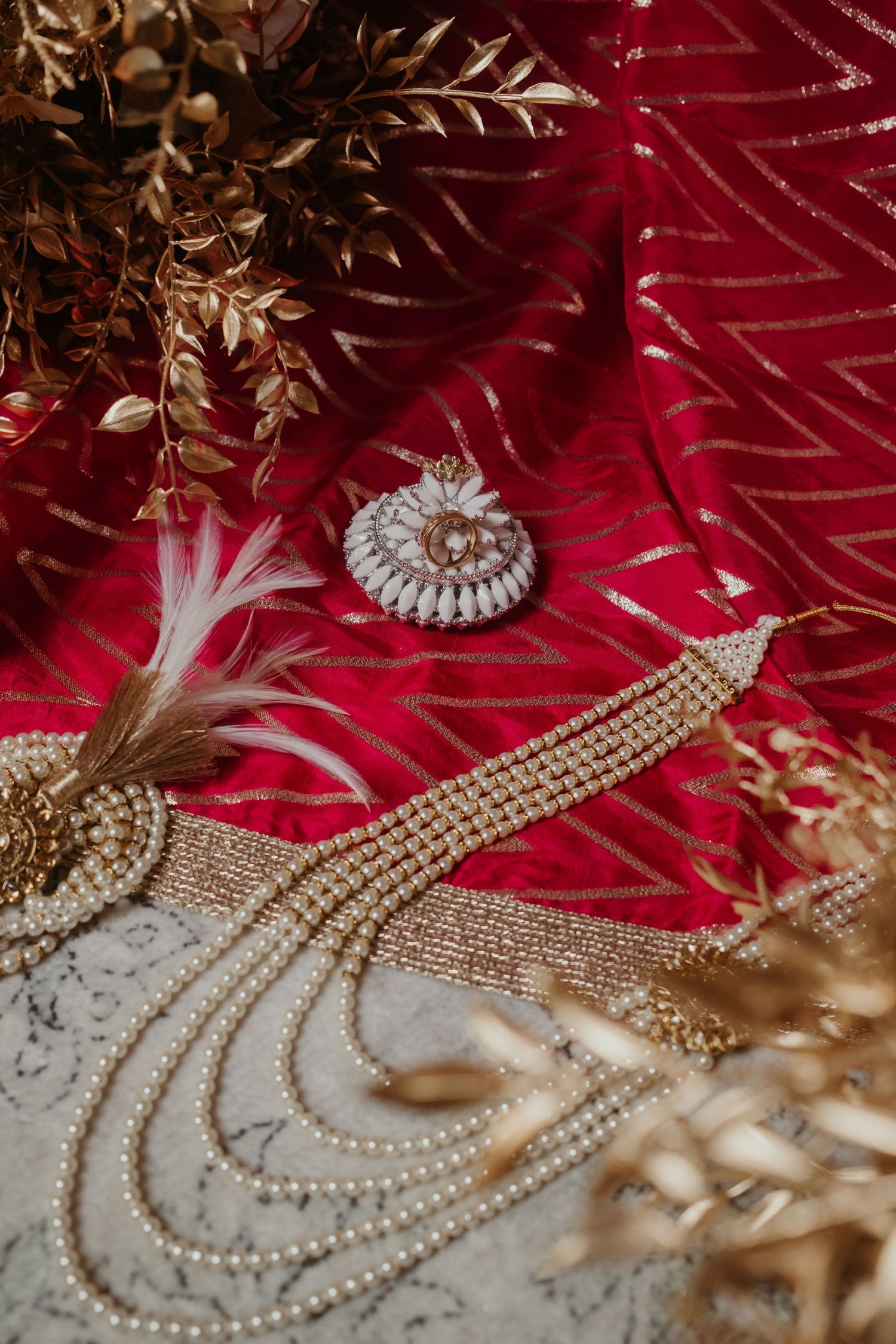 Elegant Indian bridal jewelry with pearl necklace, white floral brooch on red gold embroidered fabric with dried flowers.