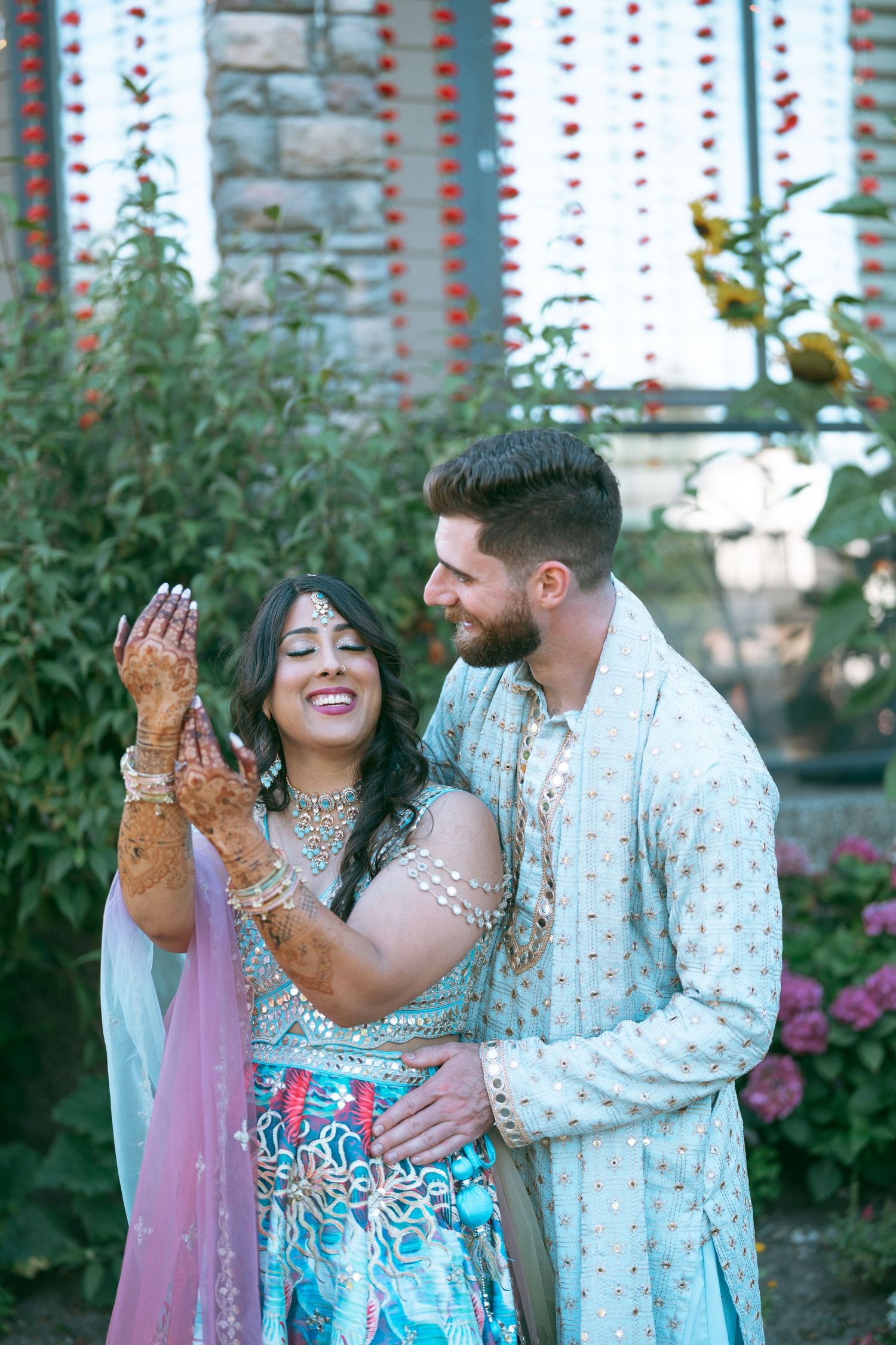 Bride in blue lehenga with mehndi hands poses with groom in light blue sherwani at outdoor Indian wedding ceremony.