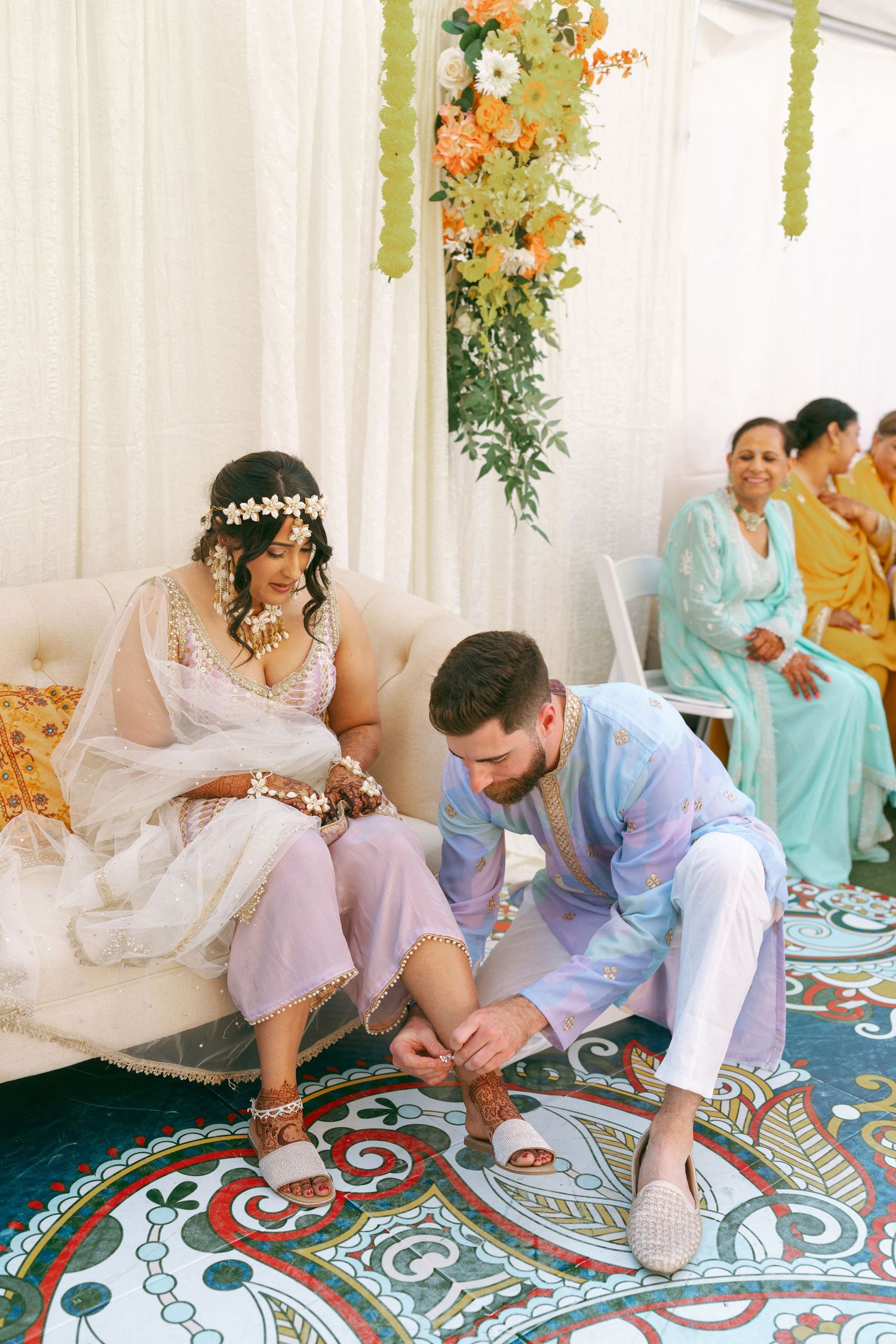 Groom in blue kurta adjusts bride's sandal during South Asian wedding ceremony on colorful mandala rug.