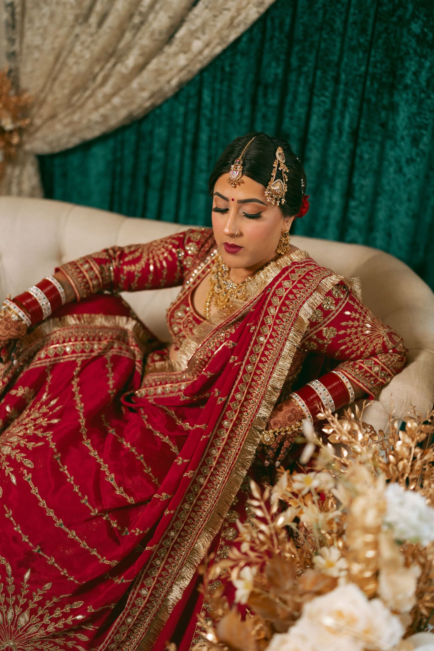 A bride in a red and gold embroidered lehenga with traditional jewelry sits gracefully on a cream sofa.