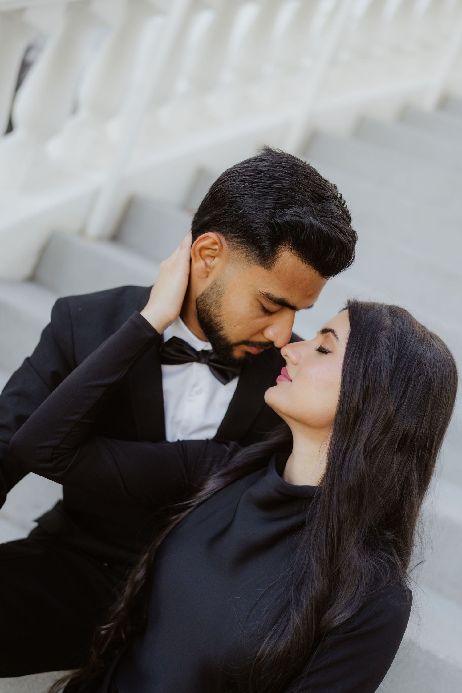 A couple in black formal attire share a romantic moment on white steps, the woman gently holding the mans face.