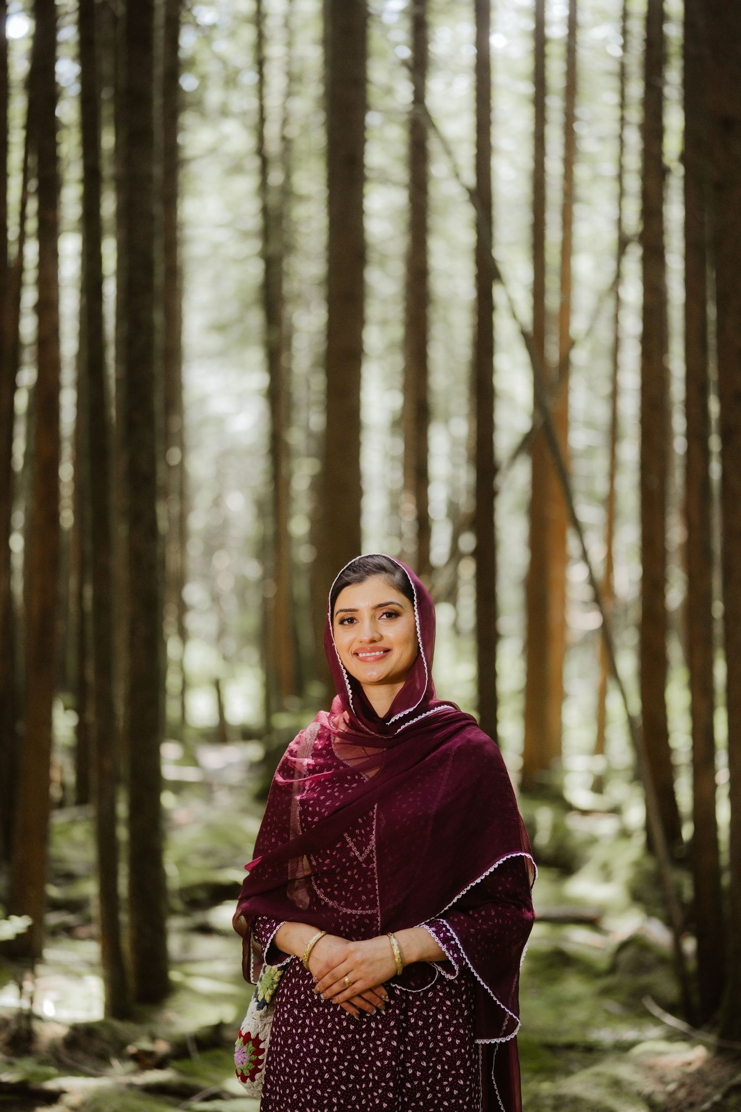 Smiling South Asian woman in burgundy hijab and traditional salwar kameez standing in a lush green forest.