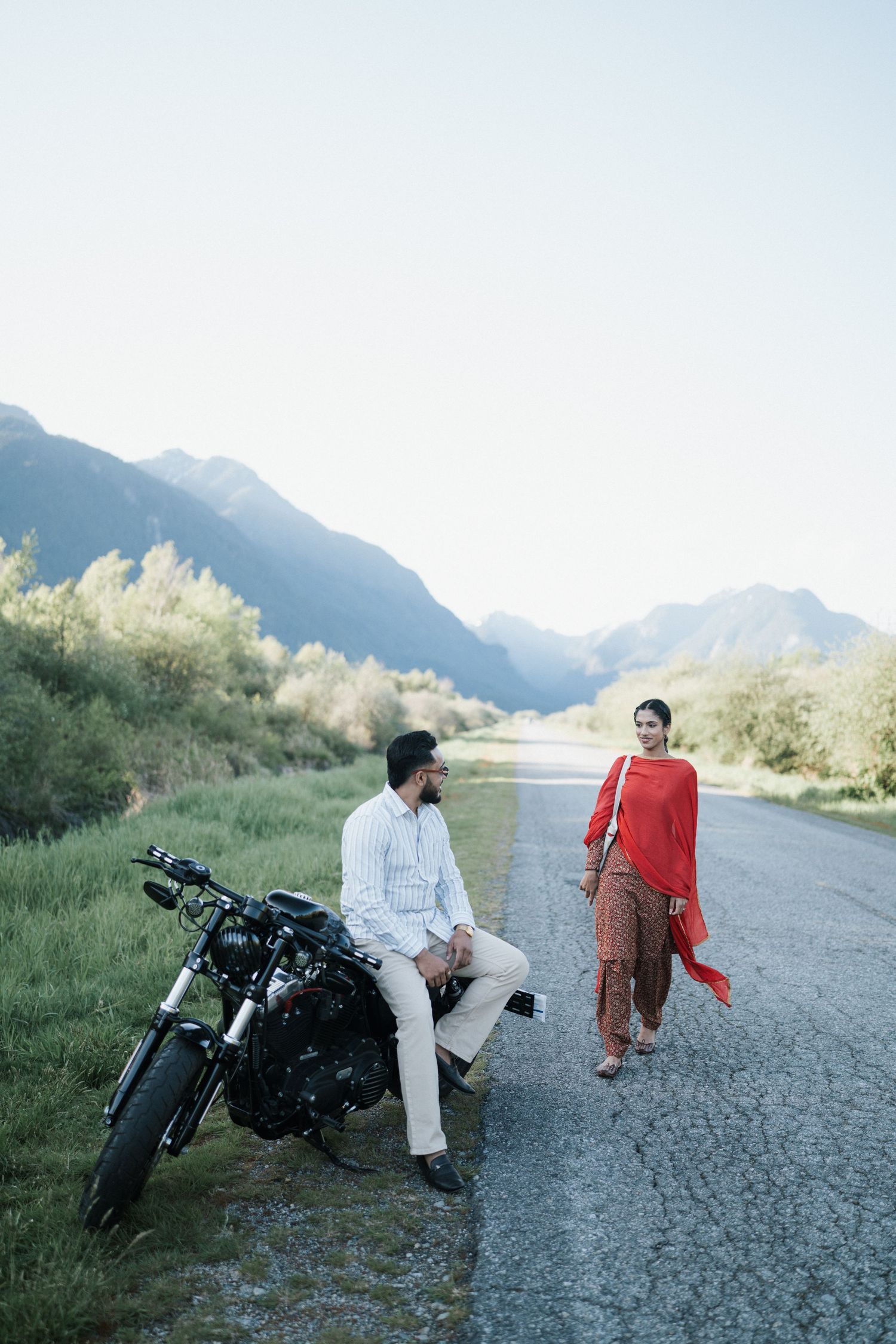 A motorcycle is parked on a gravel road with mountains in the background while two people sit and walk nearby.
