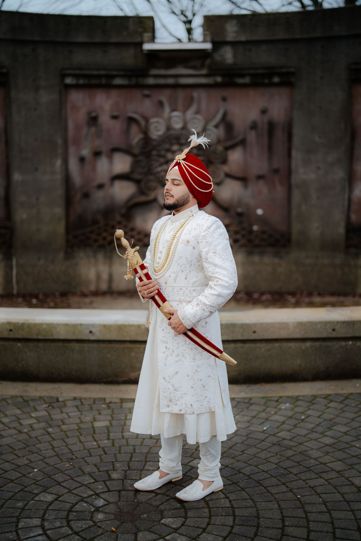Groom in white sherwani and turban poses with ceremonial sword on cobblestone ground.