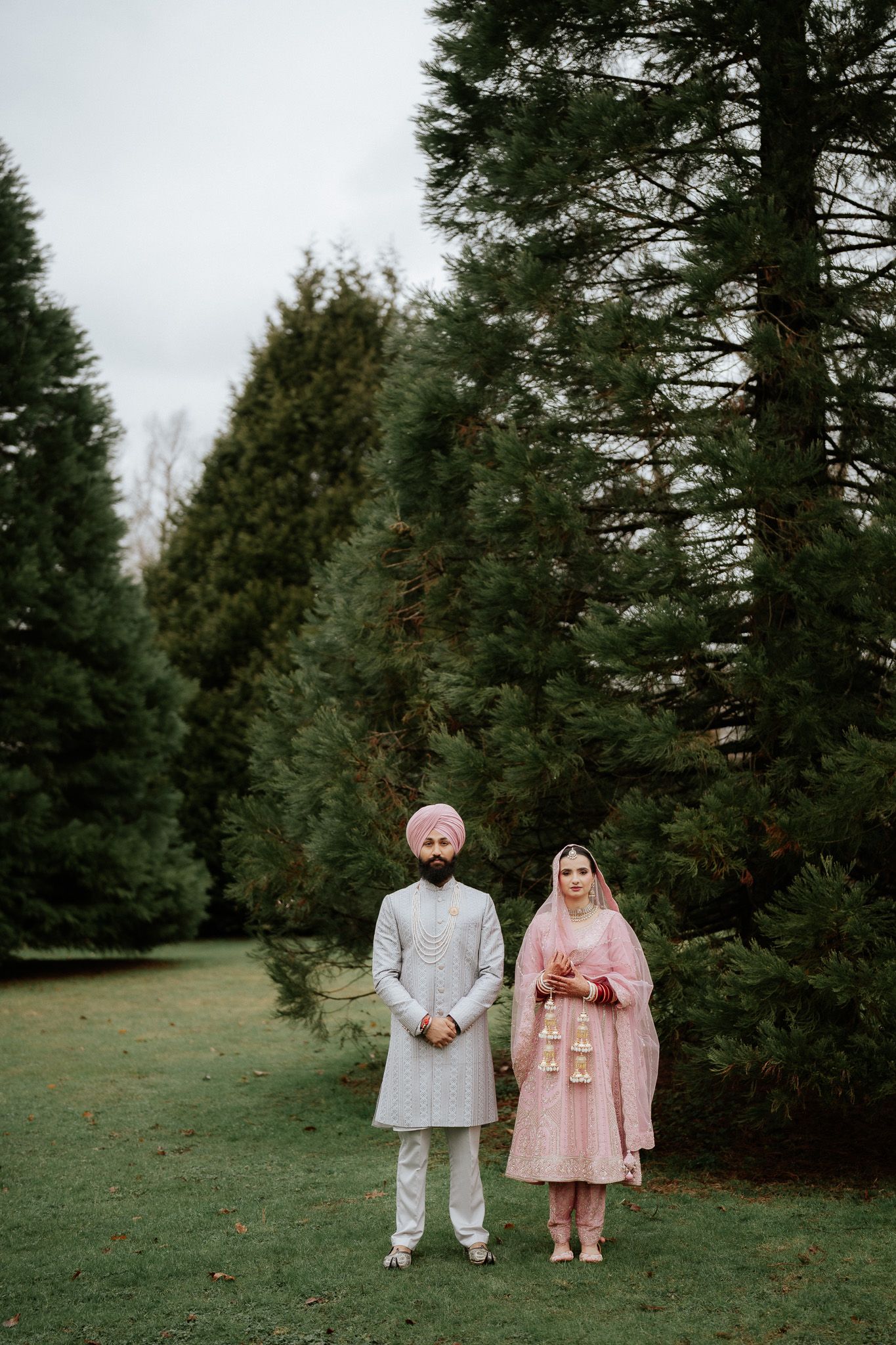 A couple stands together in traditional South Asian wedding attire against a backdrop of evergreen trees on a grassy lawn.