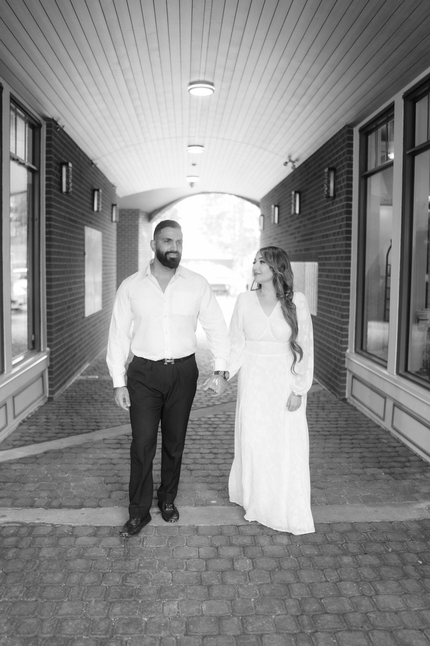 Black and white photo of a couple walking through an arched brick hallway.