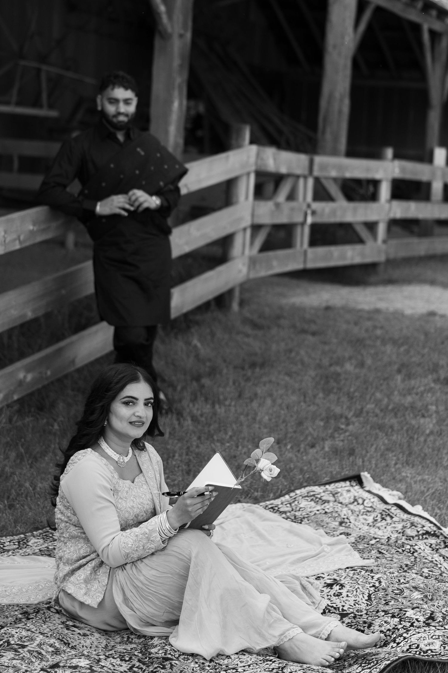 A black and white photo of a couple relaxing on a floral blanket near a wooden fence.