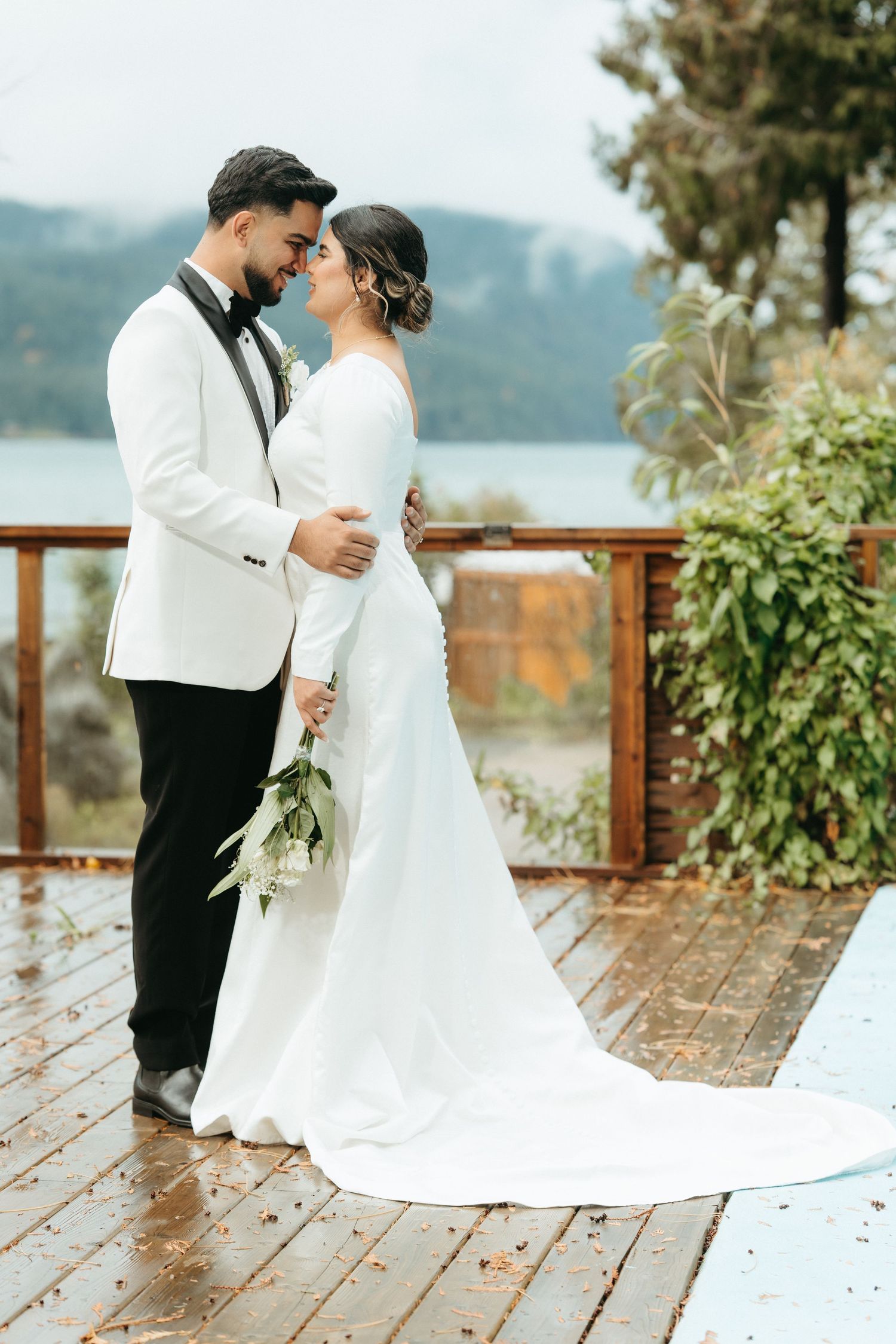 A couple in elegant wedding attire embrace on a wet wooden deck overlooking a scenic mountain lake view.