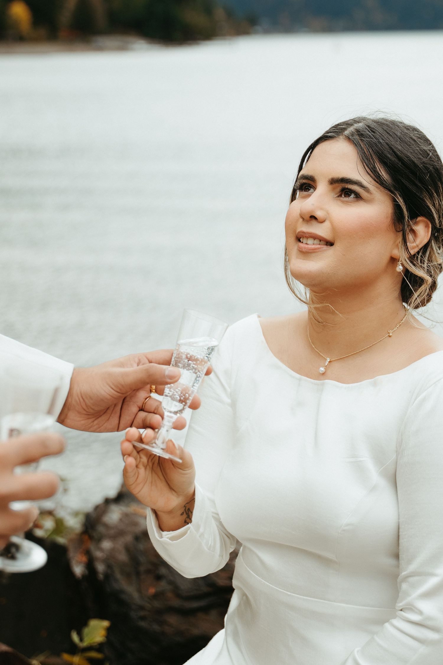 A person in a white long-sleeved dress takes a shot of liquor by a lake during a photo session.