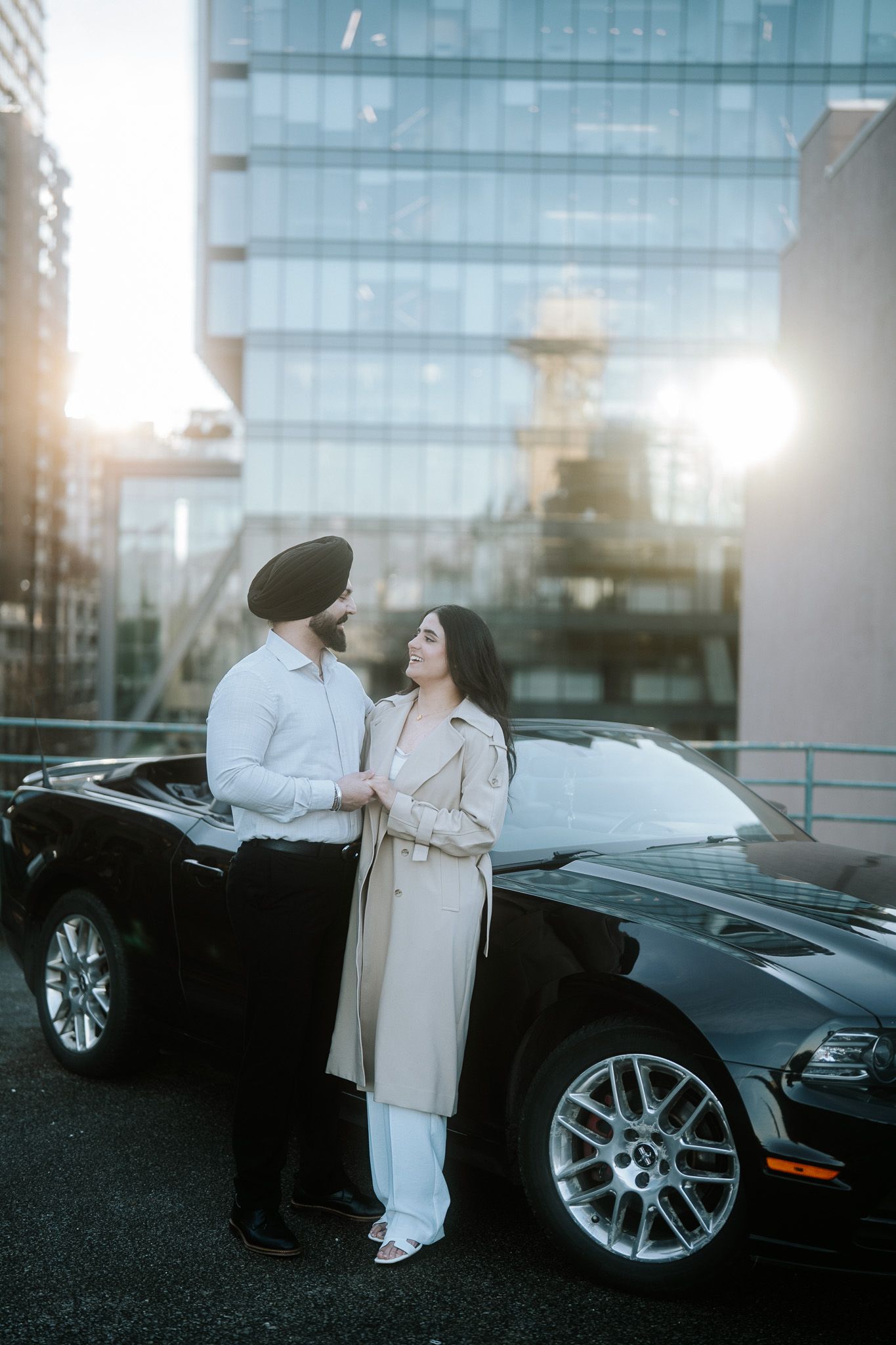 A couple stands beside a black luxury car at sunset with a modern glass building in the background.