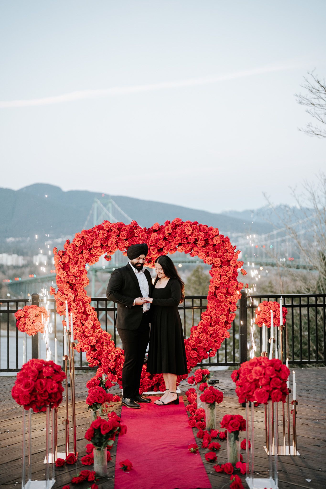 A romantic outdoor wedding ceremony setup with bright red rose arches and a red carpet against a mountain backdrop.