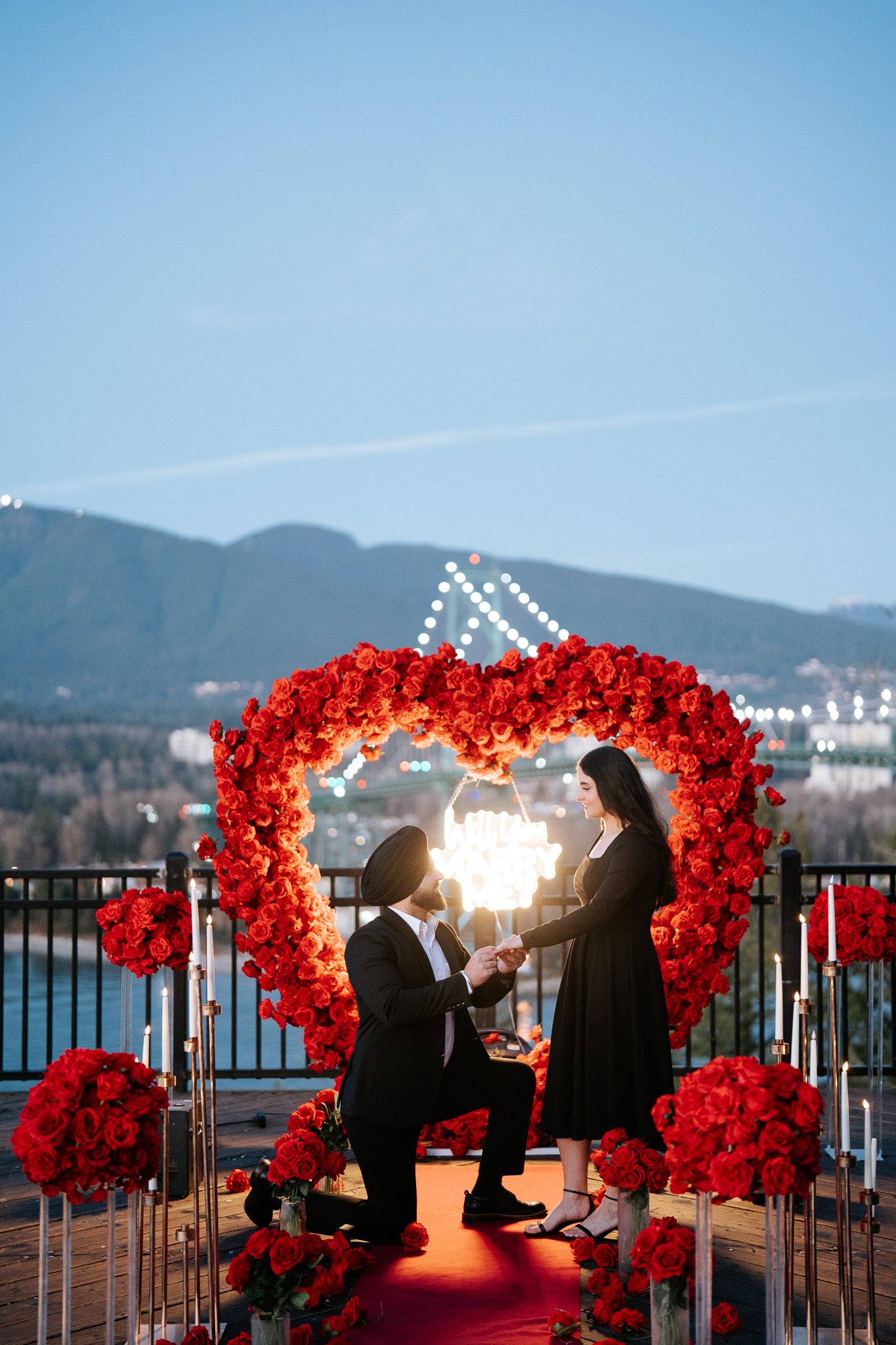 Romantic proposal setup with heart-shaped red floral arch and red carpet on balcony overlooking mountain landscape at sunset.