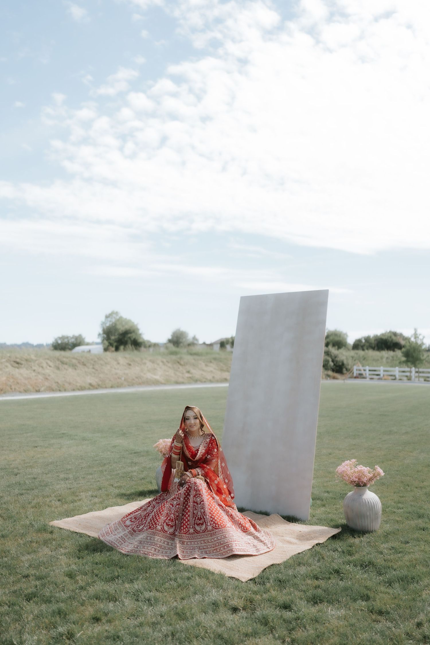Bride in red lehenga sitting on mat outdoors near white backdrop and floral vase on green lawn.