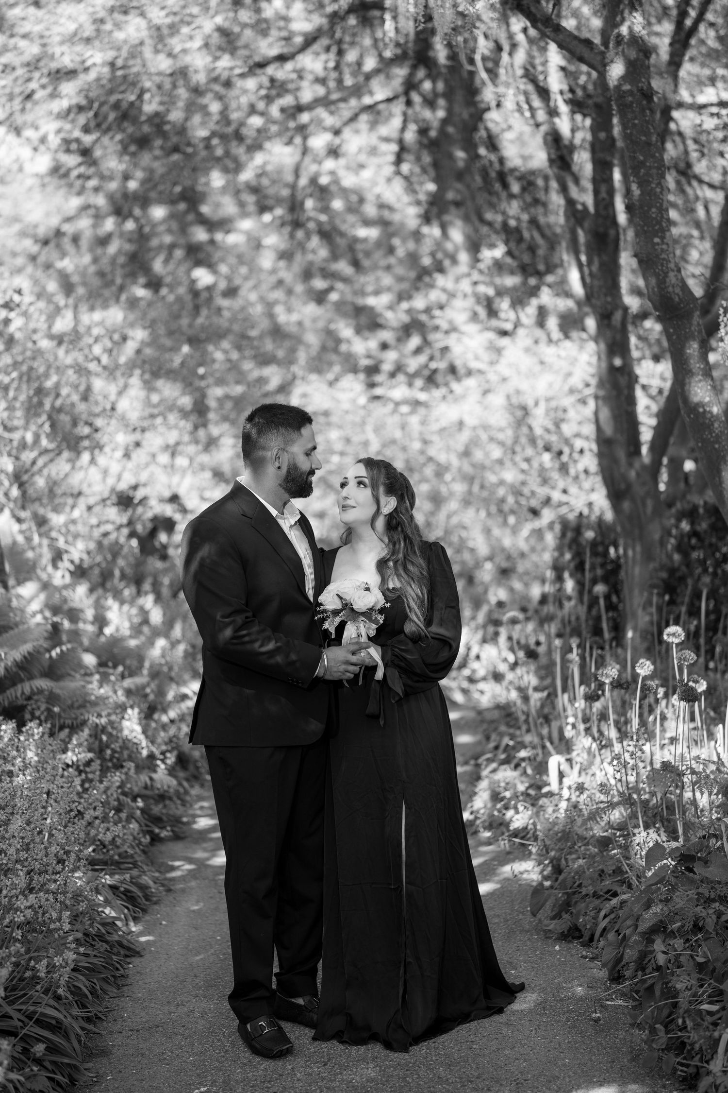 Black and white photo of a couple in formal attire embracing on a garden path surrounded by lush trees and flowers.
