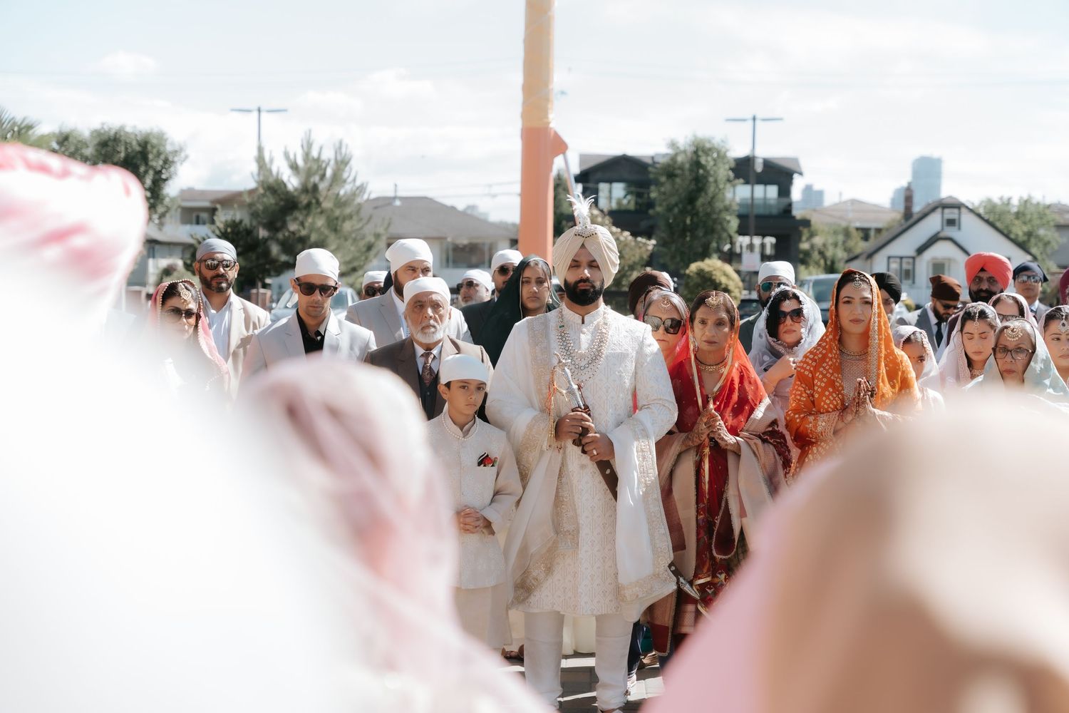 Sikh groom in white sherwani walks with family during outdoor wedding ceremony with guests in traditional attire.