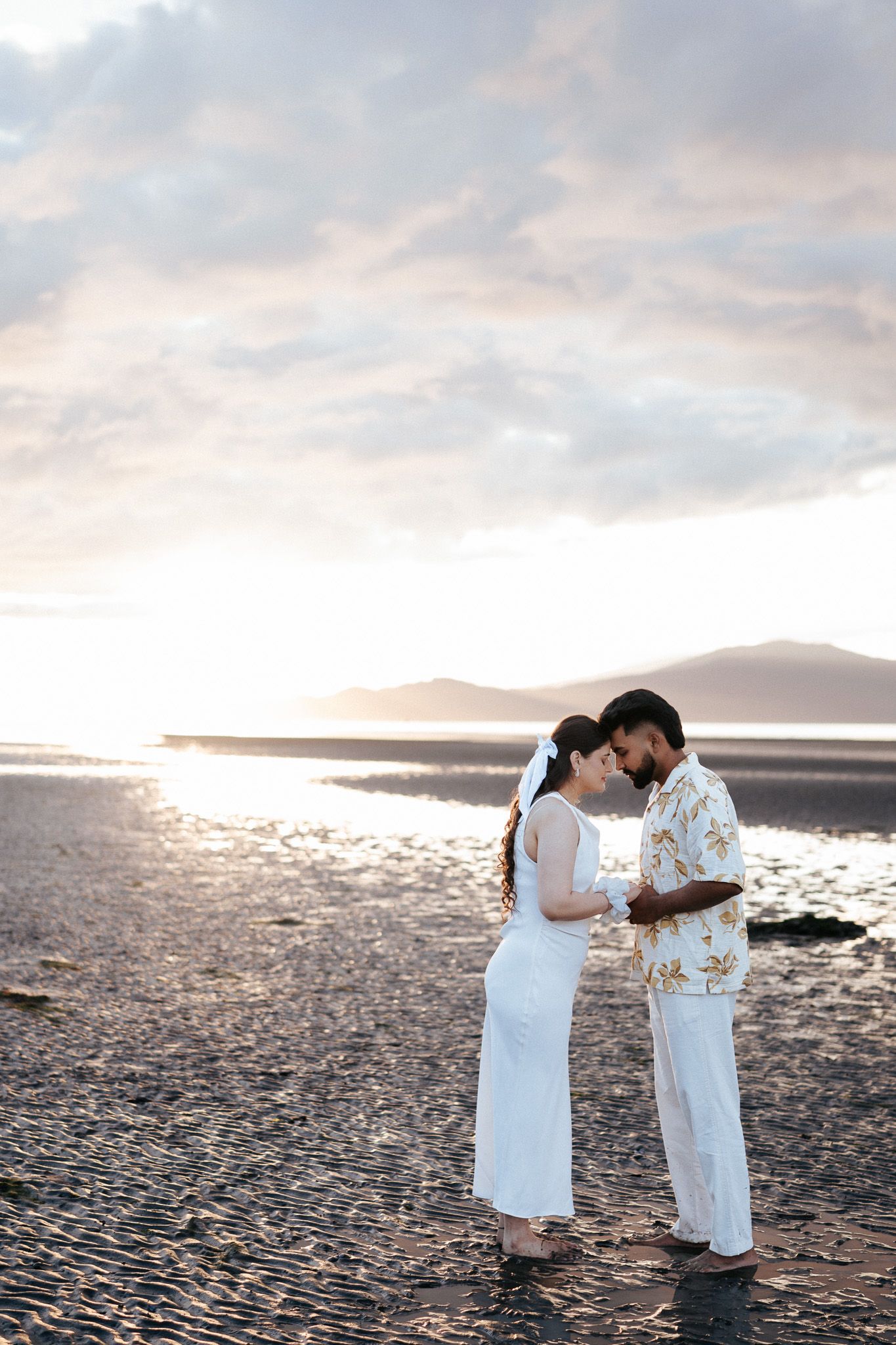 Romantic couple in white attire sharing an intimate moment on a beach at sunset with mountains behind.