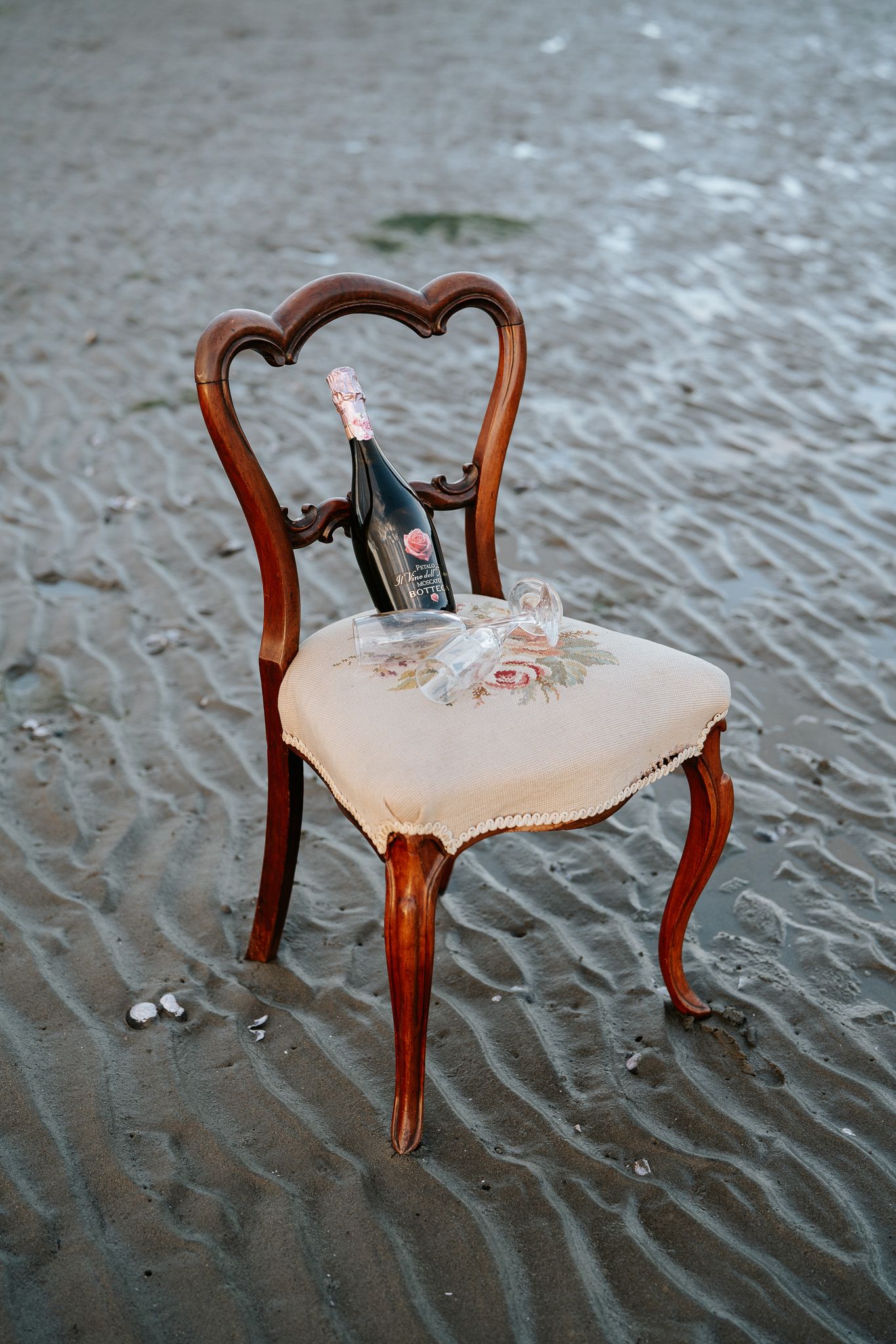An ornate vintage wooden chair with a champagne bottle and glasses sits on a rippled sandy beach.