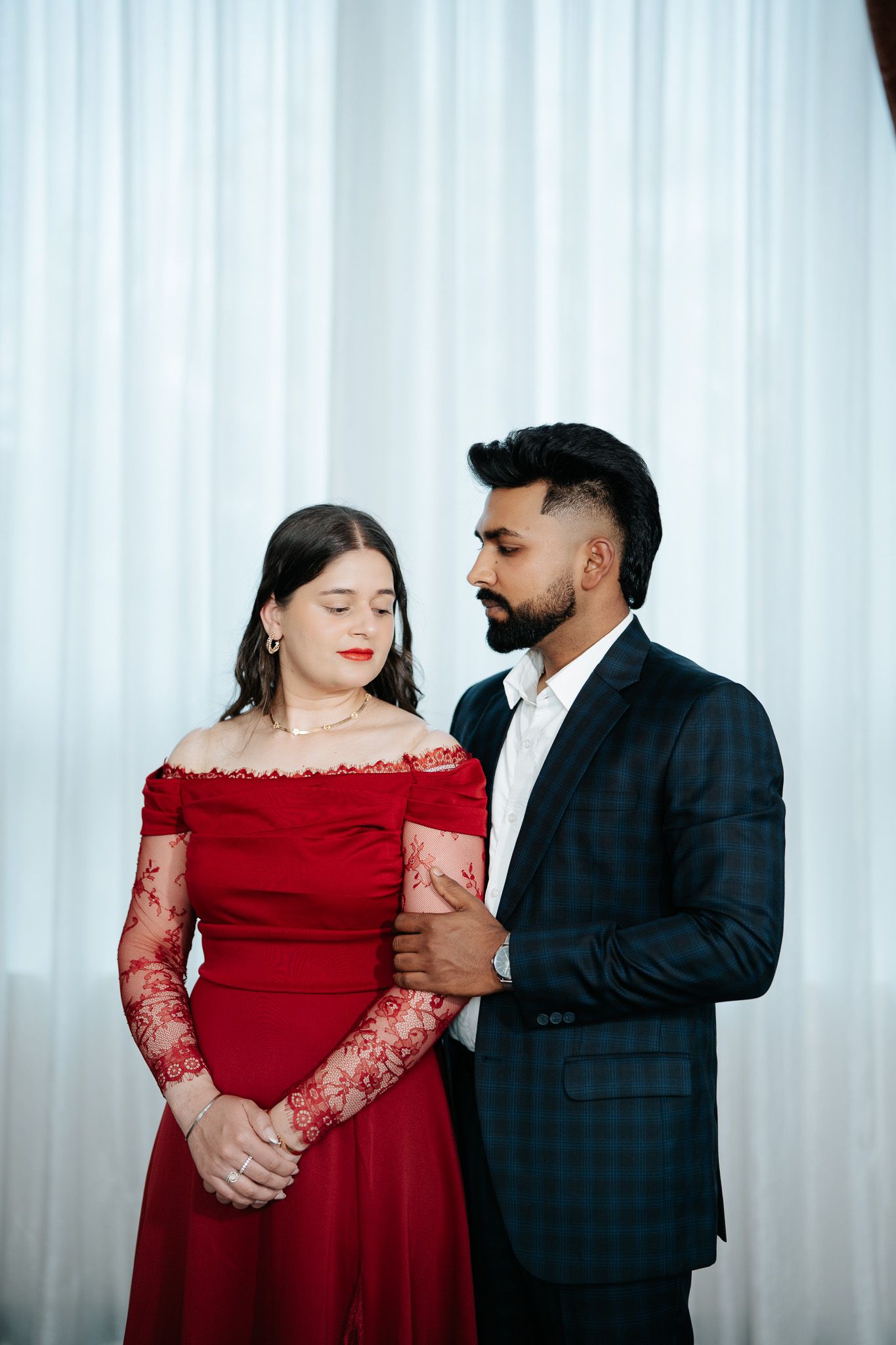Couple in elegant attire, woman in red off-shoulder gown and man in dark suit, posing together indoors.