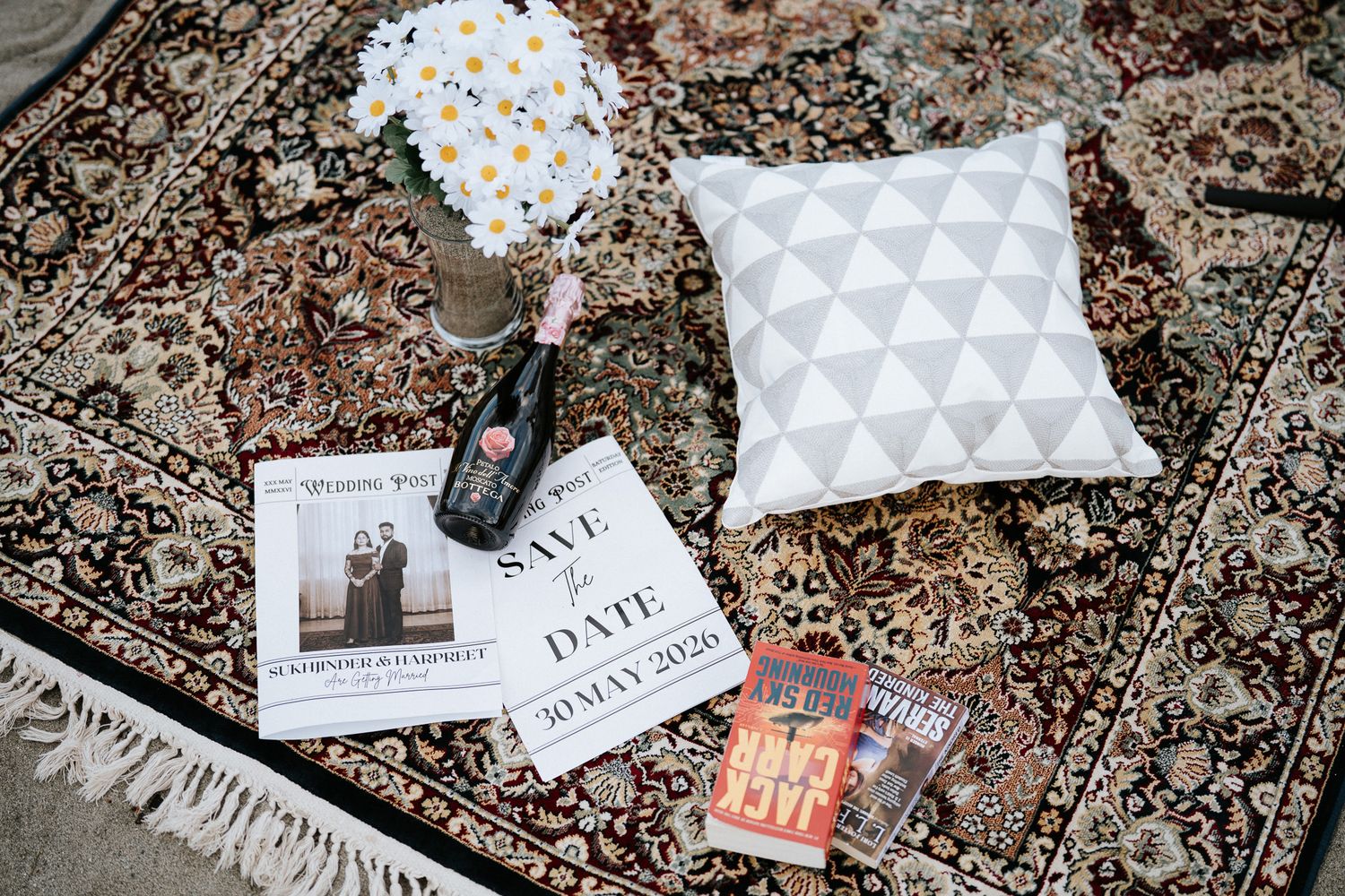 Save the date card with champagne bottle, white pillow, and daisy flowers on ornate Persian rug.