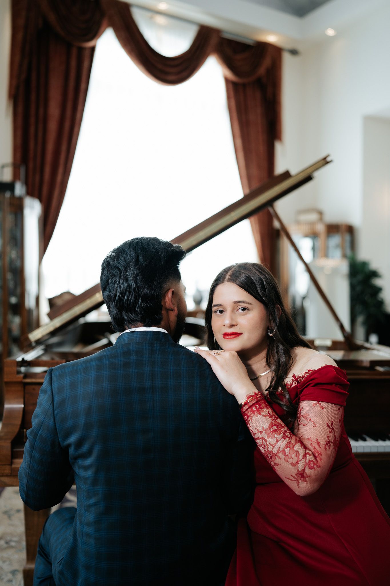 Woman in red off-shoulder gown gazing at partner near a grand piano with burgundy drapes backdrop.