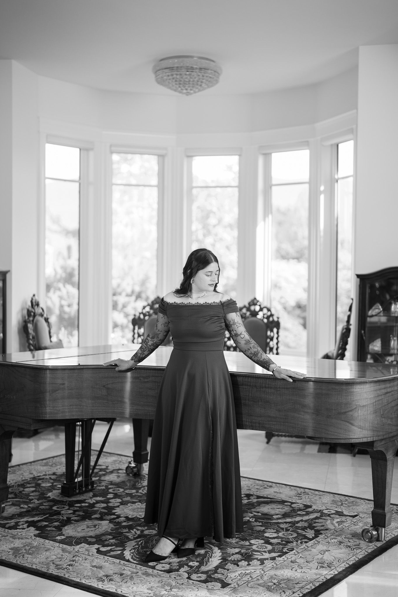 Elegant woman in black off-shoulder gown stands beside a grand piano in bright white room, black and white photo.