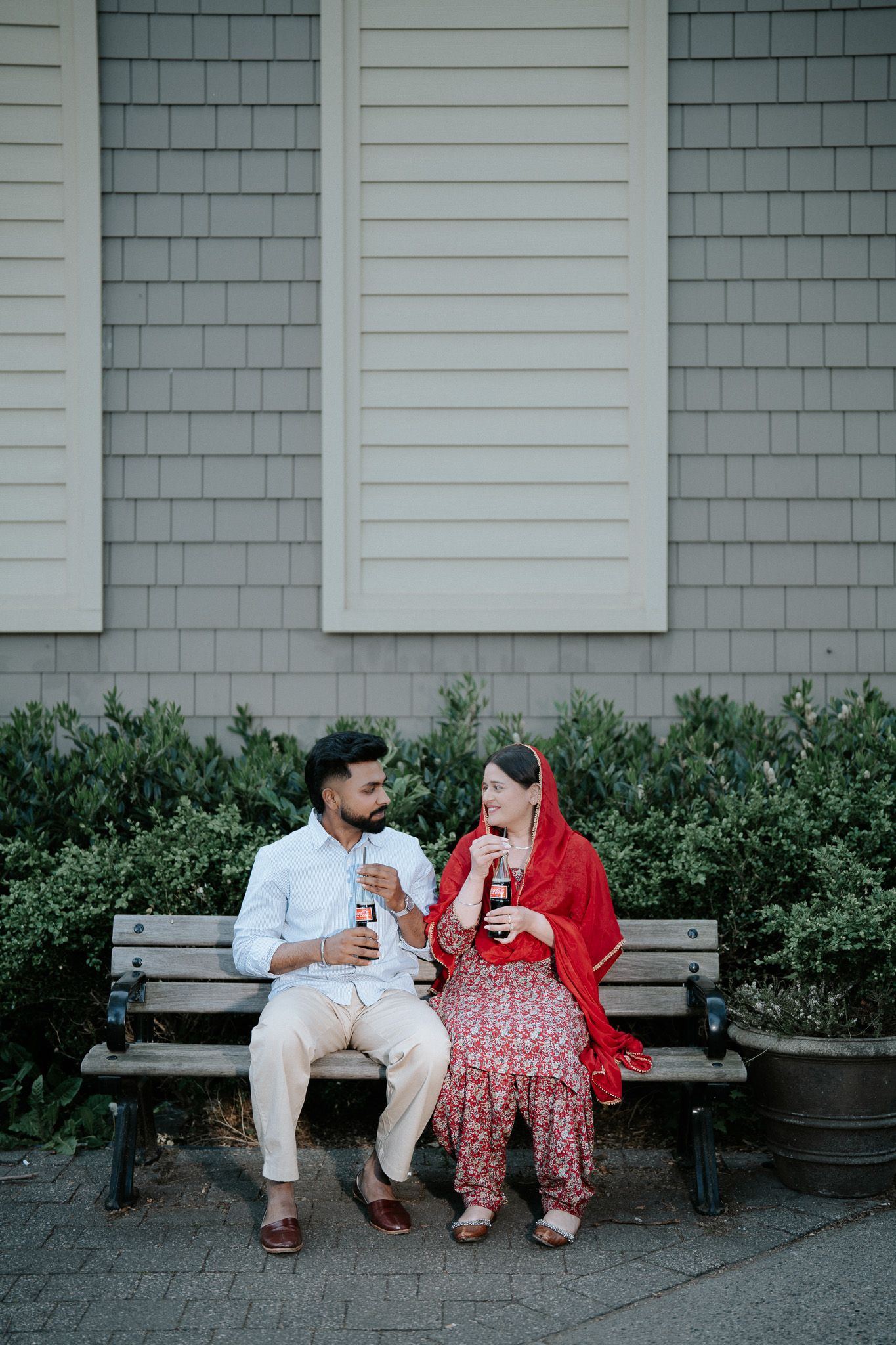 Couple in traditional South Asian attire sitting on a bench, sharing drinks and smiling at each other outdoors.