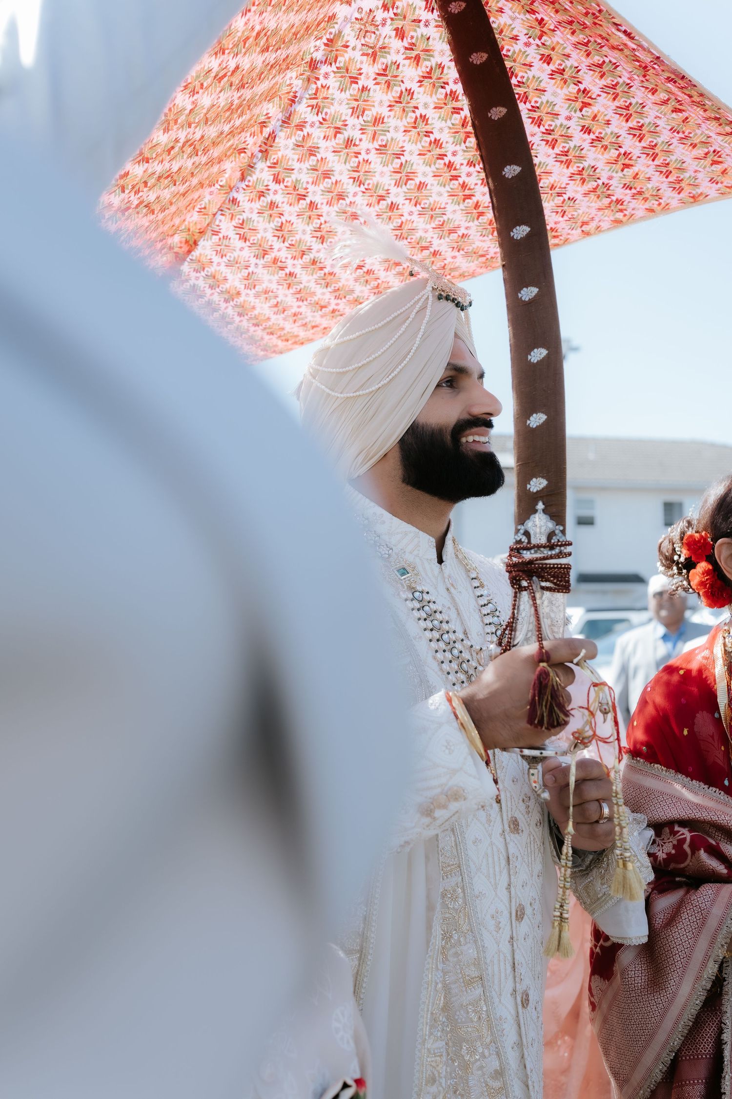 Sikh groom in white sherwani and turban smiling under decorative red umbrella during traditional wedding ceremony.