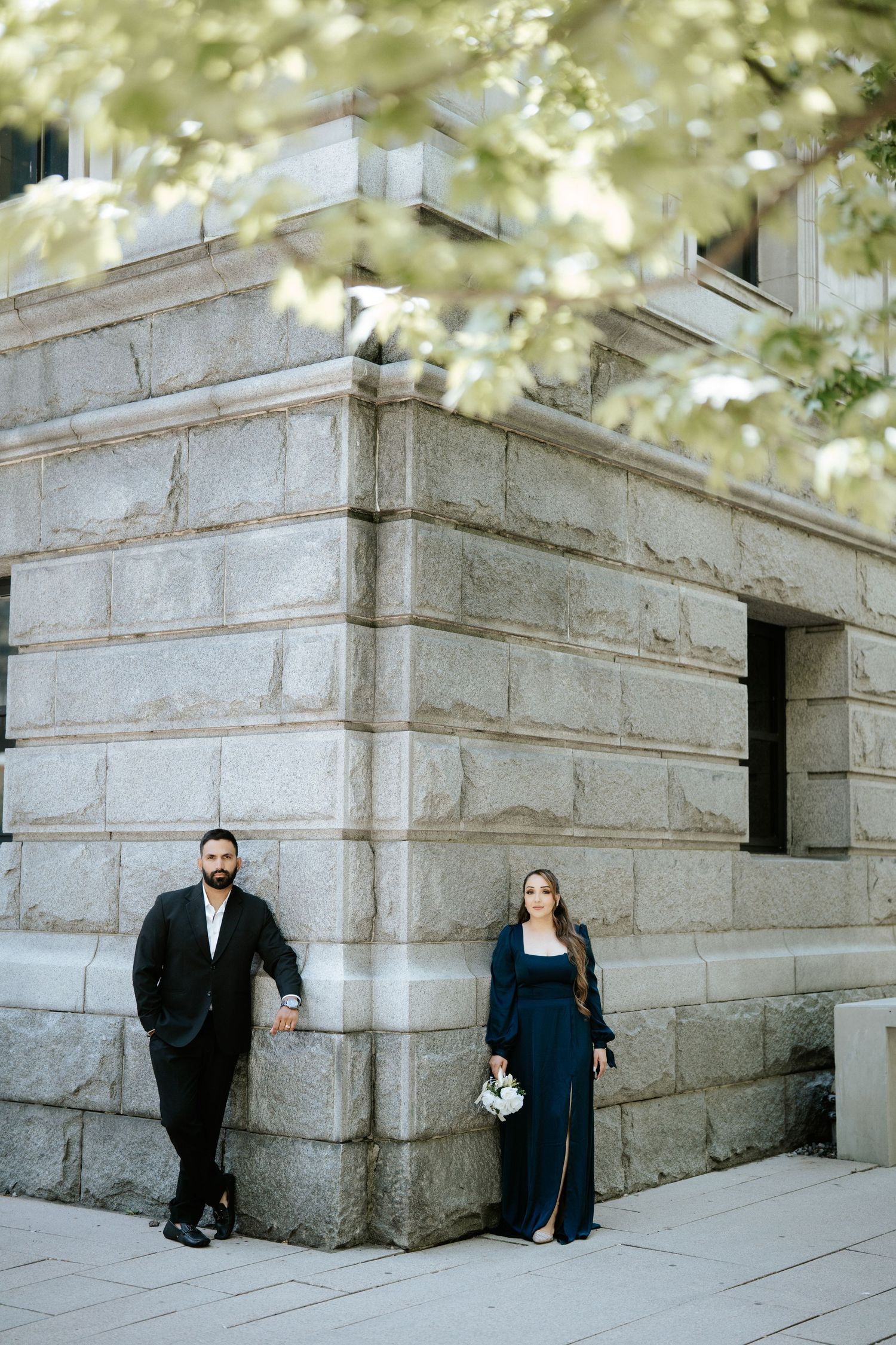Elegant couple posing by a grand stone building corner, man in black suit and woman in navy dress holding flowers.