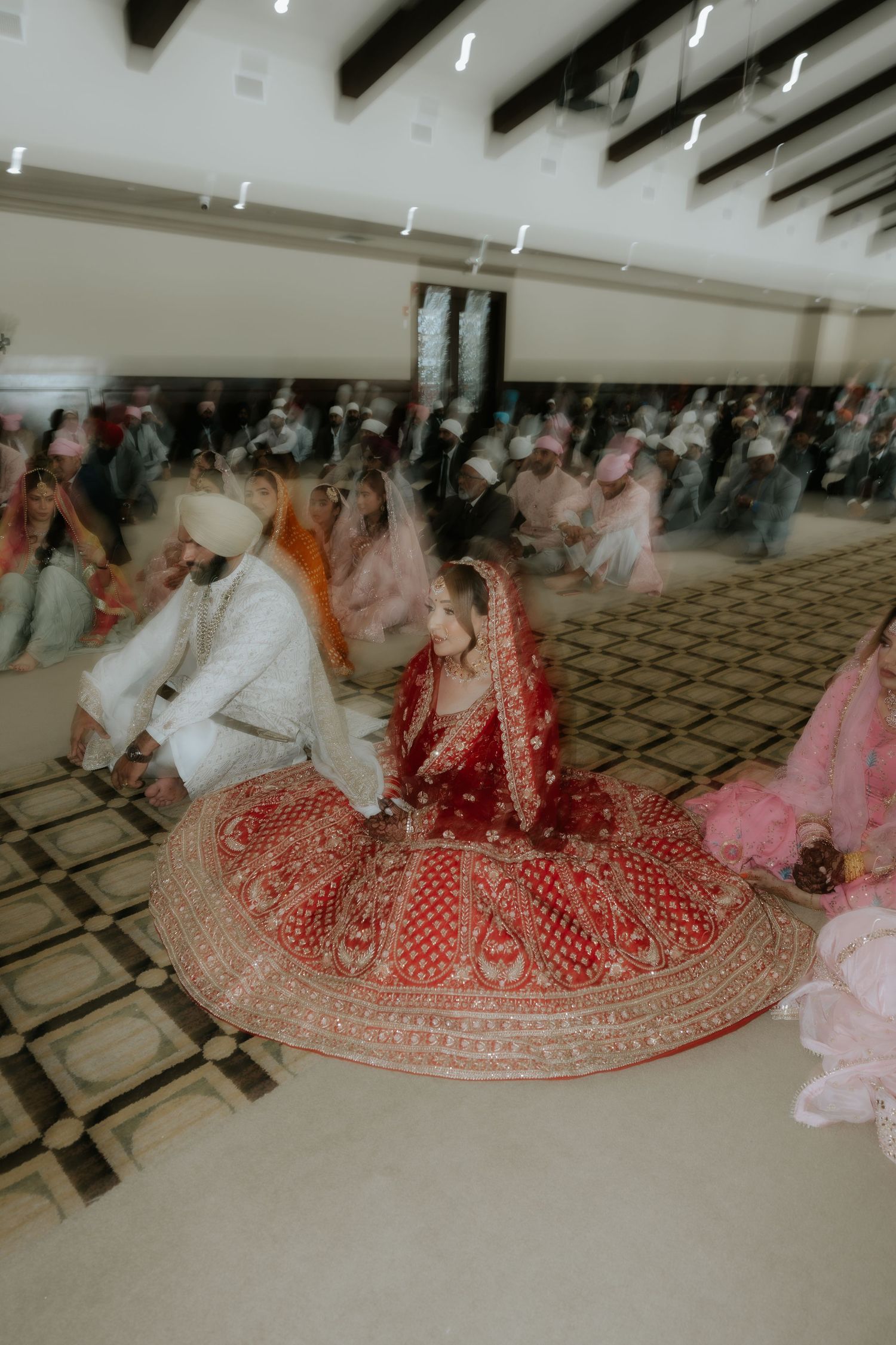 Sikh bride in red lehenga and groom in white sherwani seated during traditional Anand Karaj wedding ceremony.