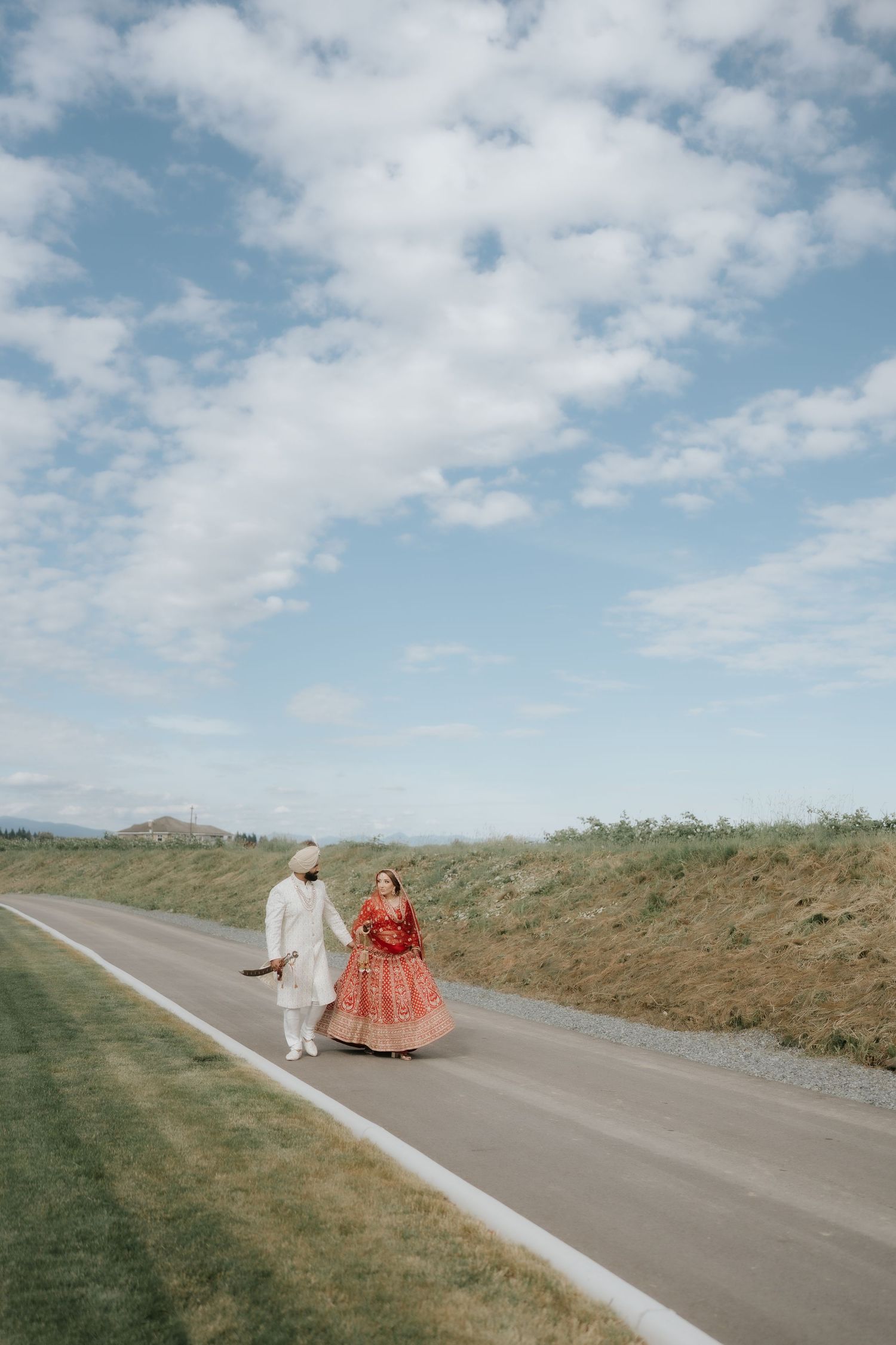 Indian bride in red lehenga and groom in white sherwani walking together on a scenic road under blue sky.