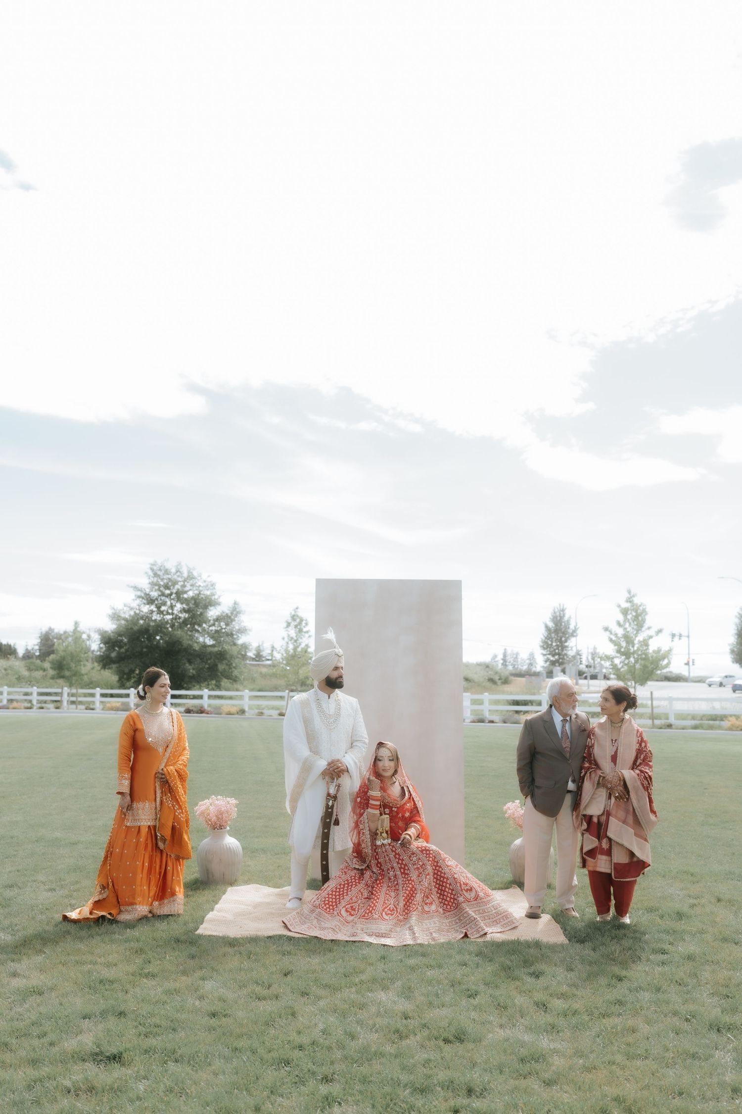 South Asian bride in red lehenga seated outdoors at wedding ceremony with family in traditional attire on green lawn.