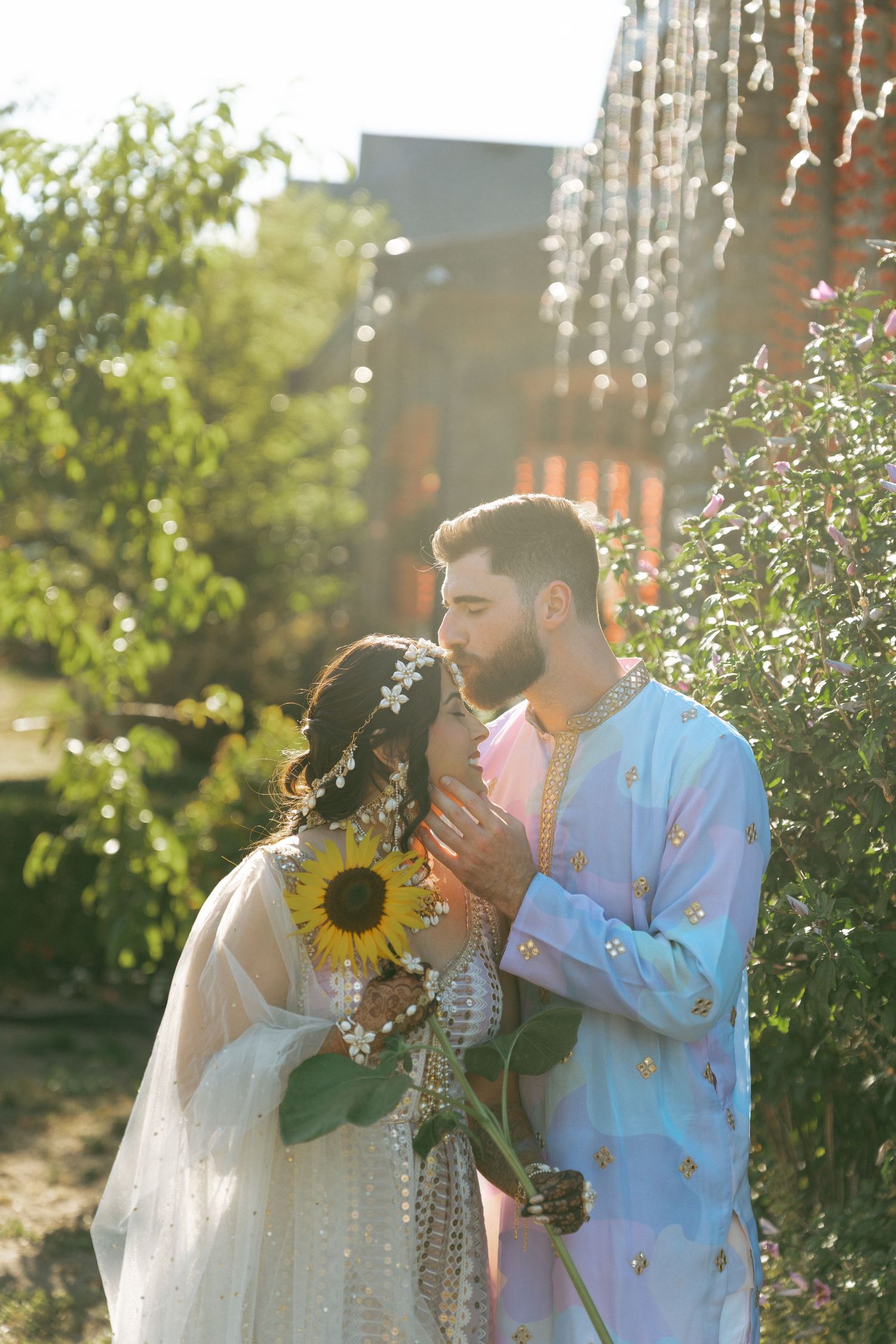 Couple in traditional South Asian attire sharing a romantic moment with a sunflower in a sunlit garden.