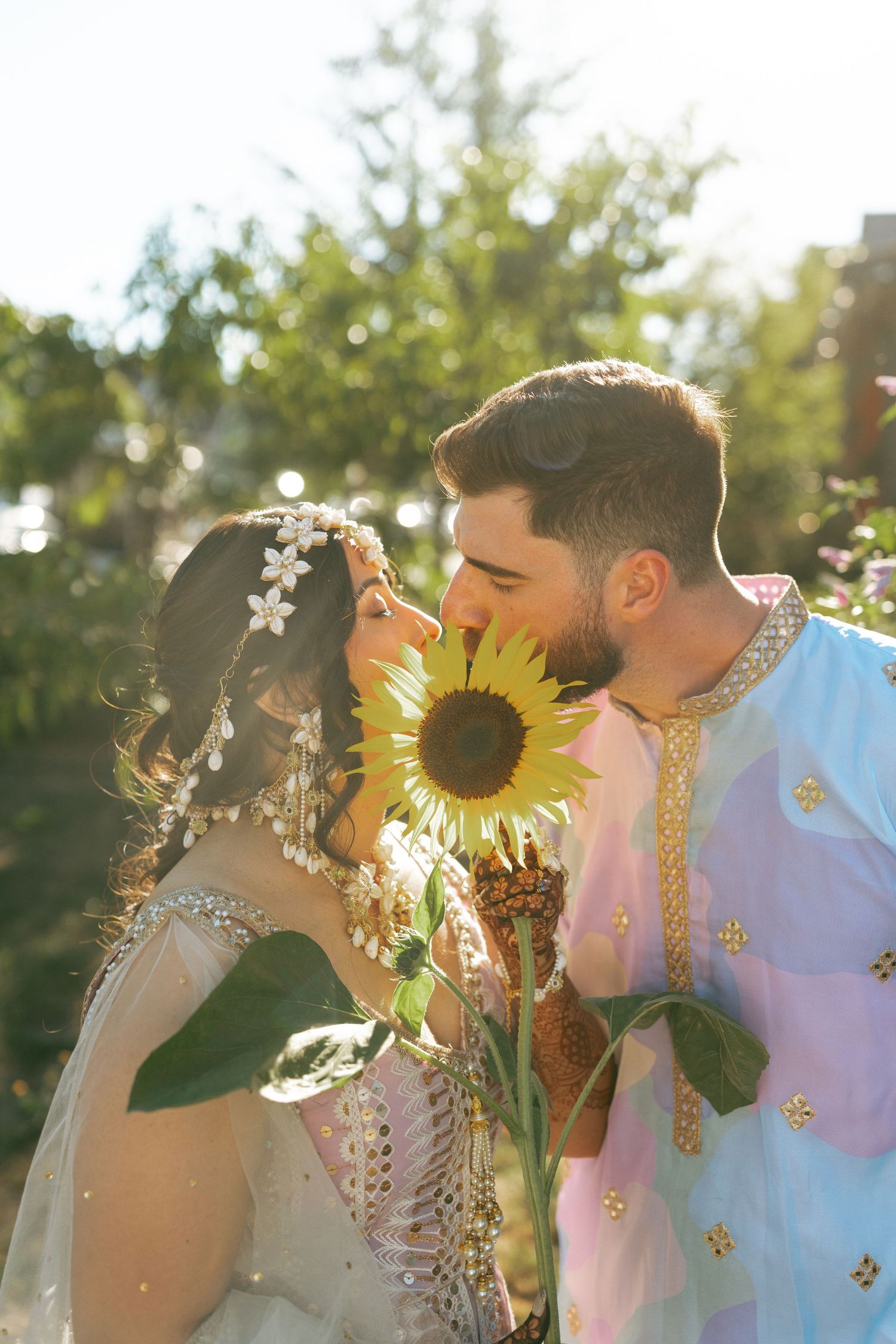Bride and groom in traditional attire sharing a romantic moment behind a sunflower in a sunlit garden.