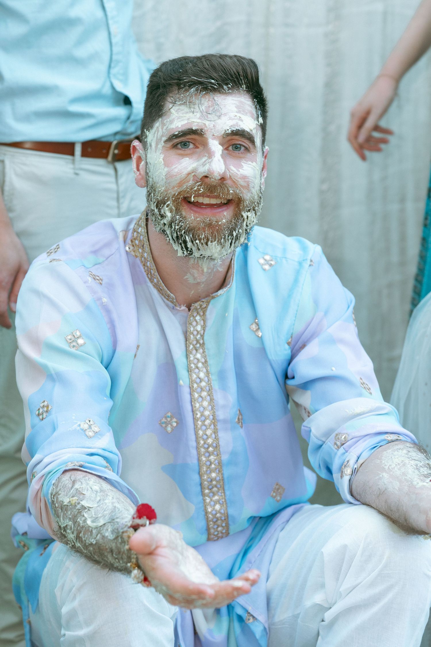 Man with white powder on face wearing light blue embroidered kurta, smiling and holding a red flower.