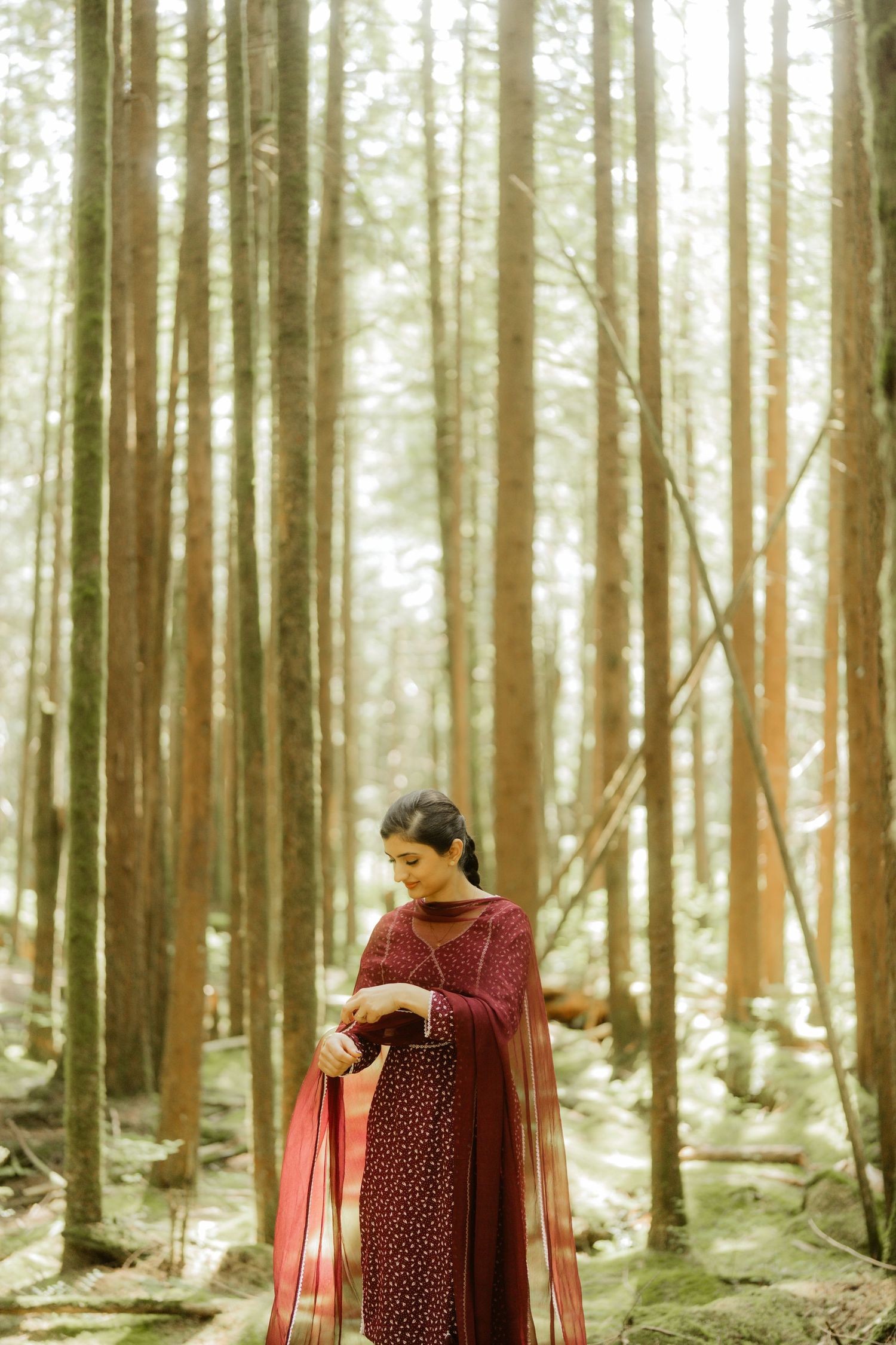 Woman in elegant burgundy Indian salwar kameez with dupatta standing gracefully among tall pine trees in a forest.