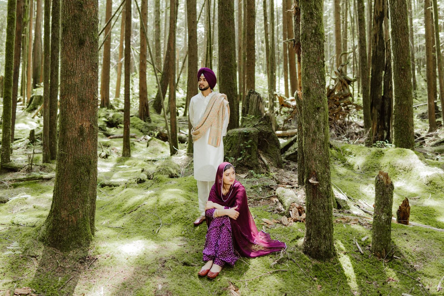 A Sikh couple poses in a lush forest, man standing in white kurta and purple turban, woman seated in maroon.