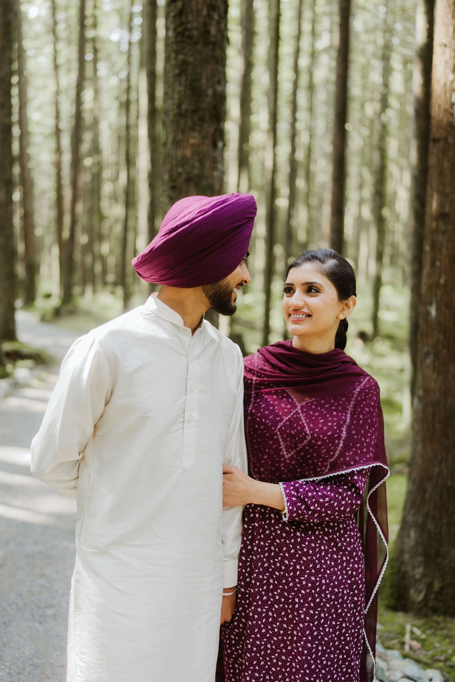 Sikh couple walking in forest, man in white kurta and purple turban, woman in maroon salwar kameez.