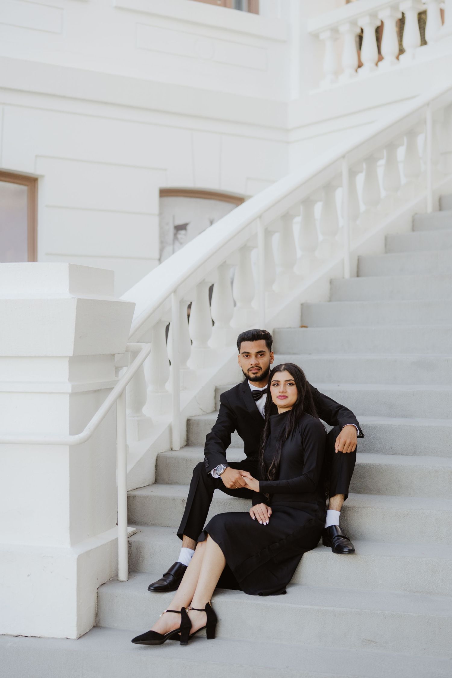 Couple in black formal attire sitting on white marble staircase steps of an elegant classical building.