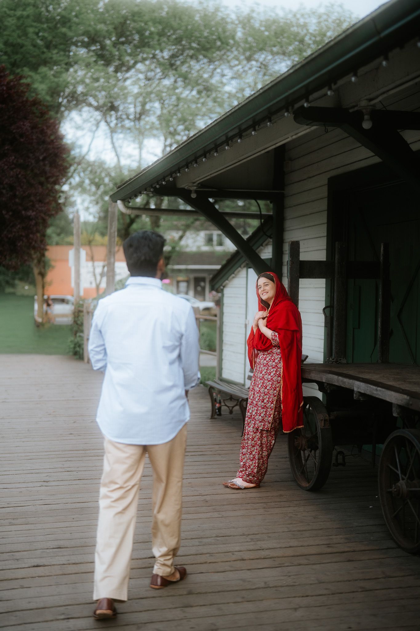 Man in light blue shirt walks toward smiling woman in red traditional attire on a wooden heritage station platform.