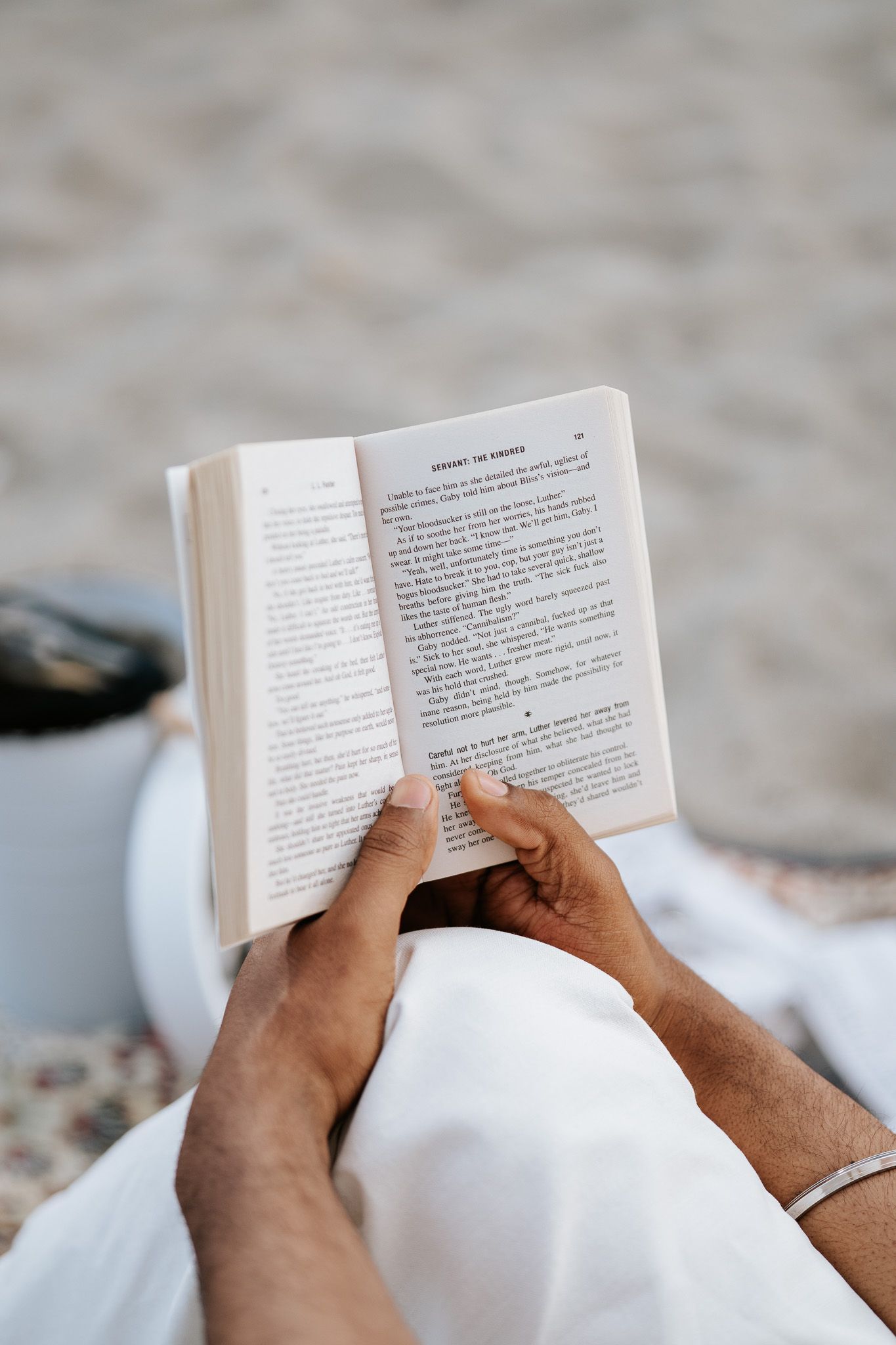 Person reading an open book on a sandy beach, wearing white clothing with a drink nearby.