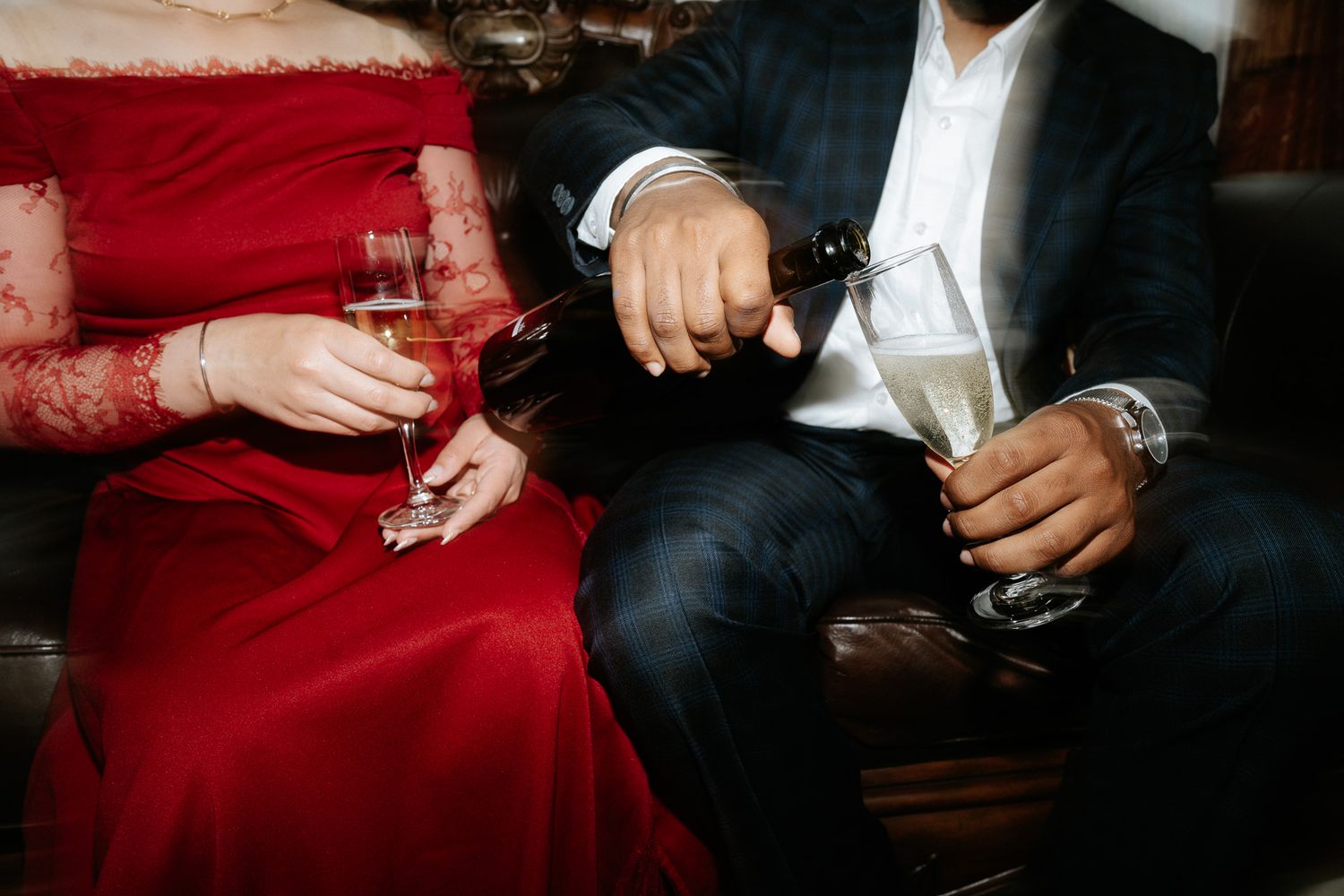 Close-up of couple in formal wear pouring champagne into glasses while seated on a sofa.
