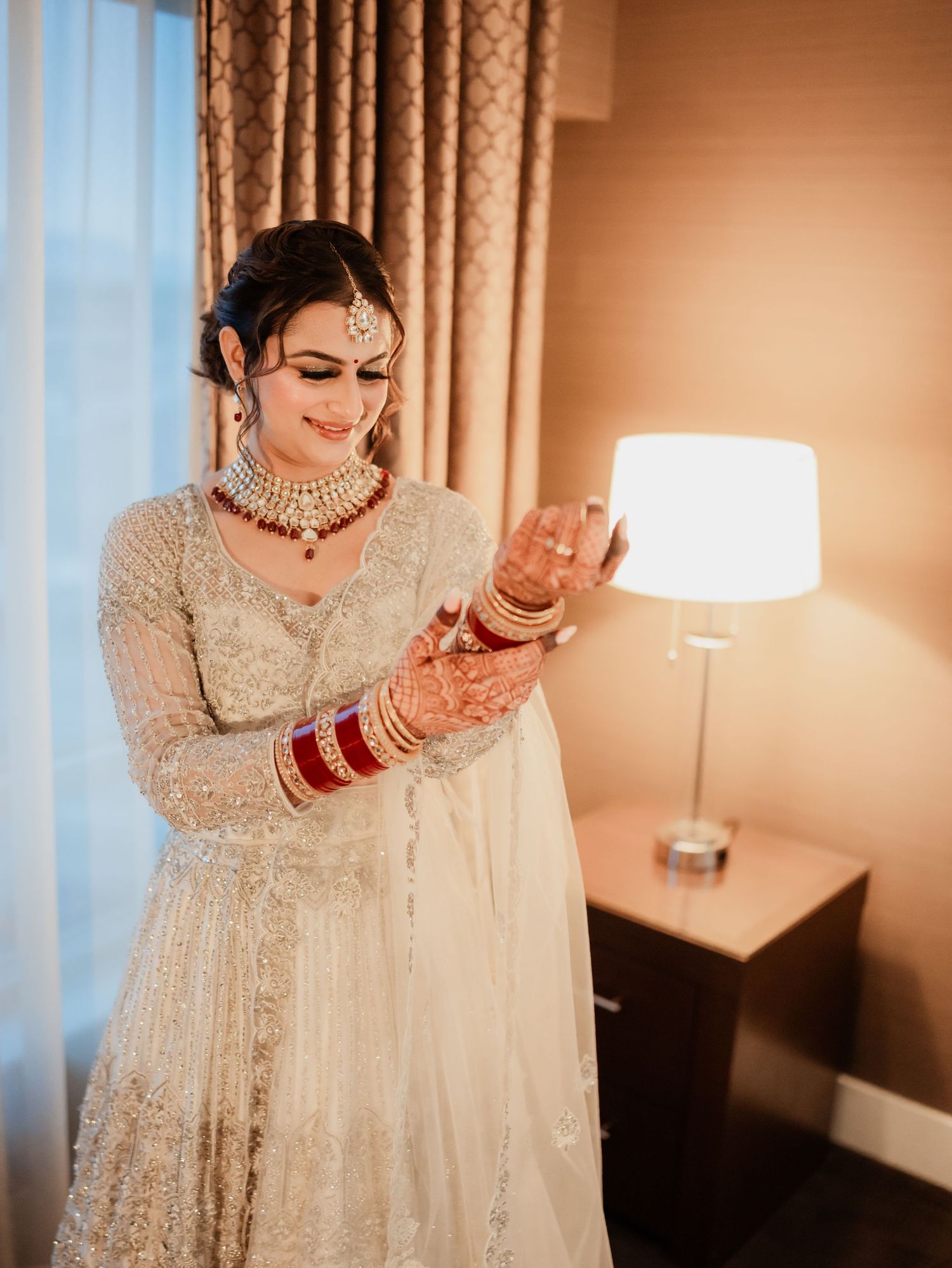 A bride in an elegant white lehenga poses gracefully in a hotel room with warm lighting.