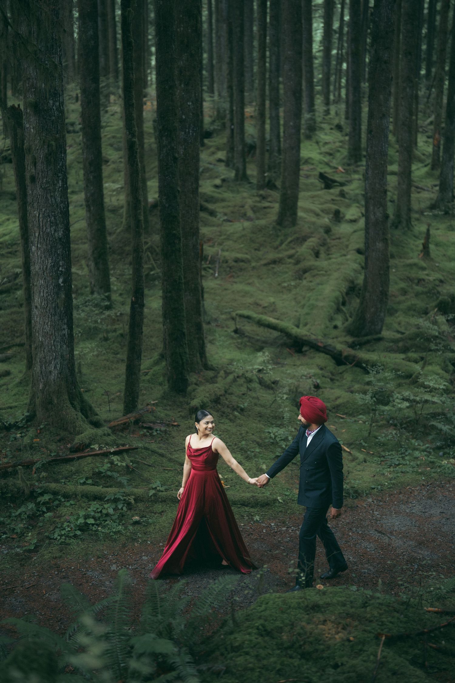 Couple in red dress and black suit holds hands while walking through a misty forest.