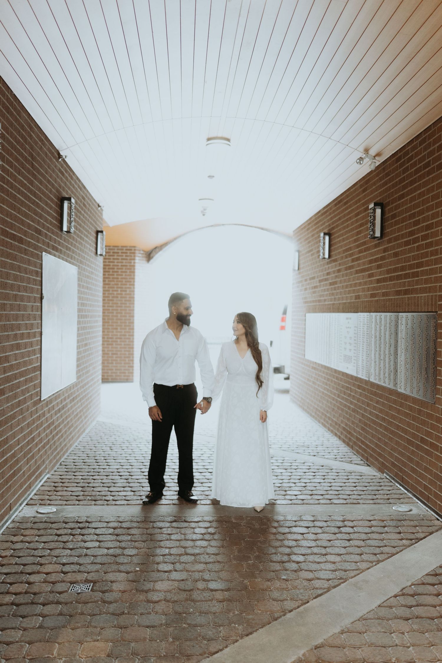 A couple stands in silhouette against natural light in a brick corridor.