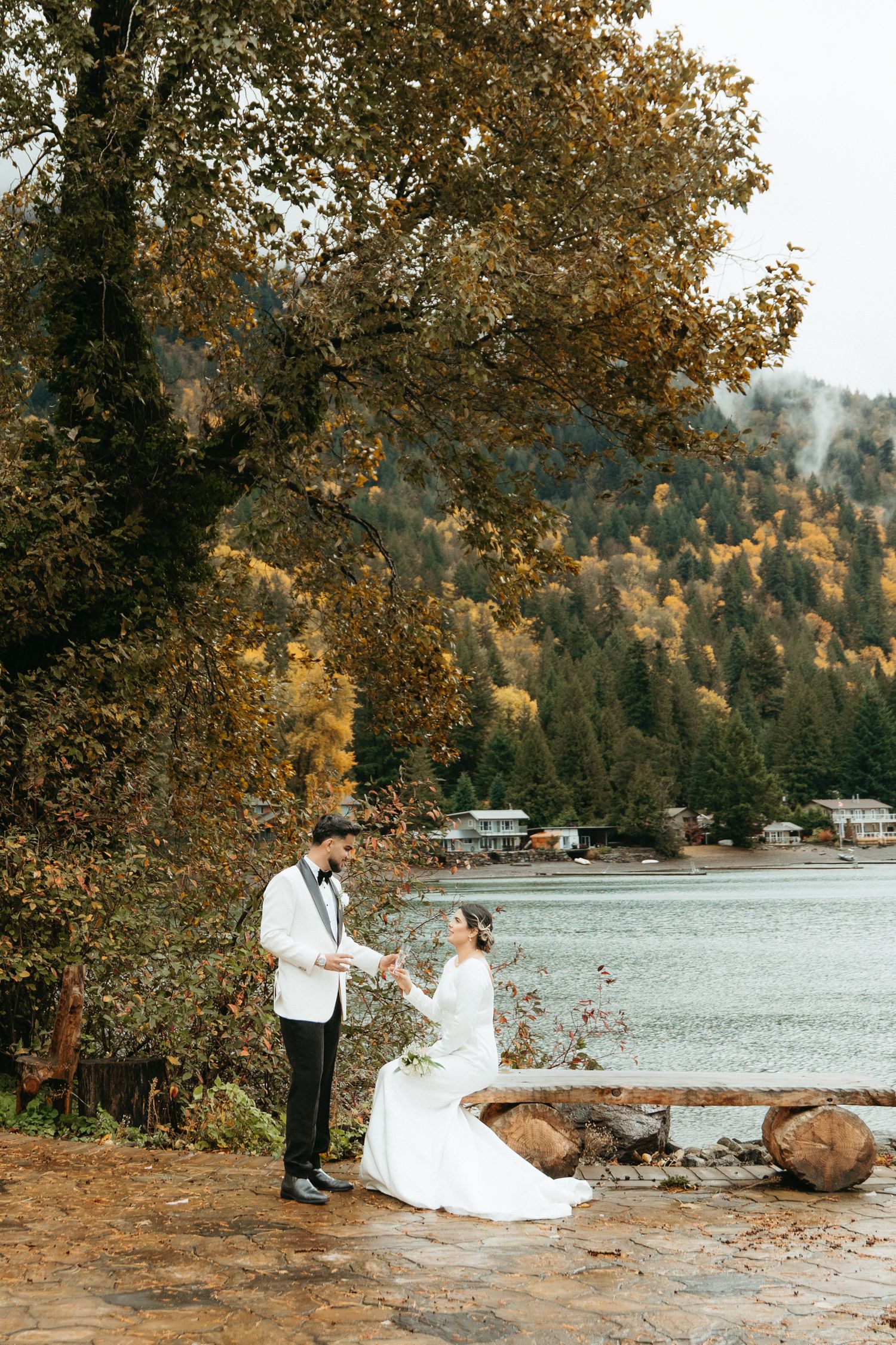 A couple shares a romantic moment by a lake in autumn with golden trees and waterfront cabins in the background.
