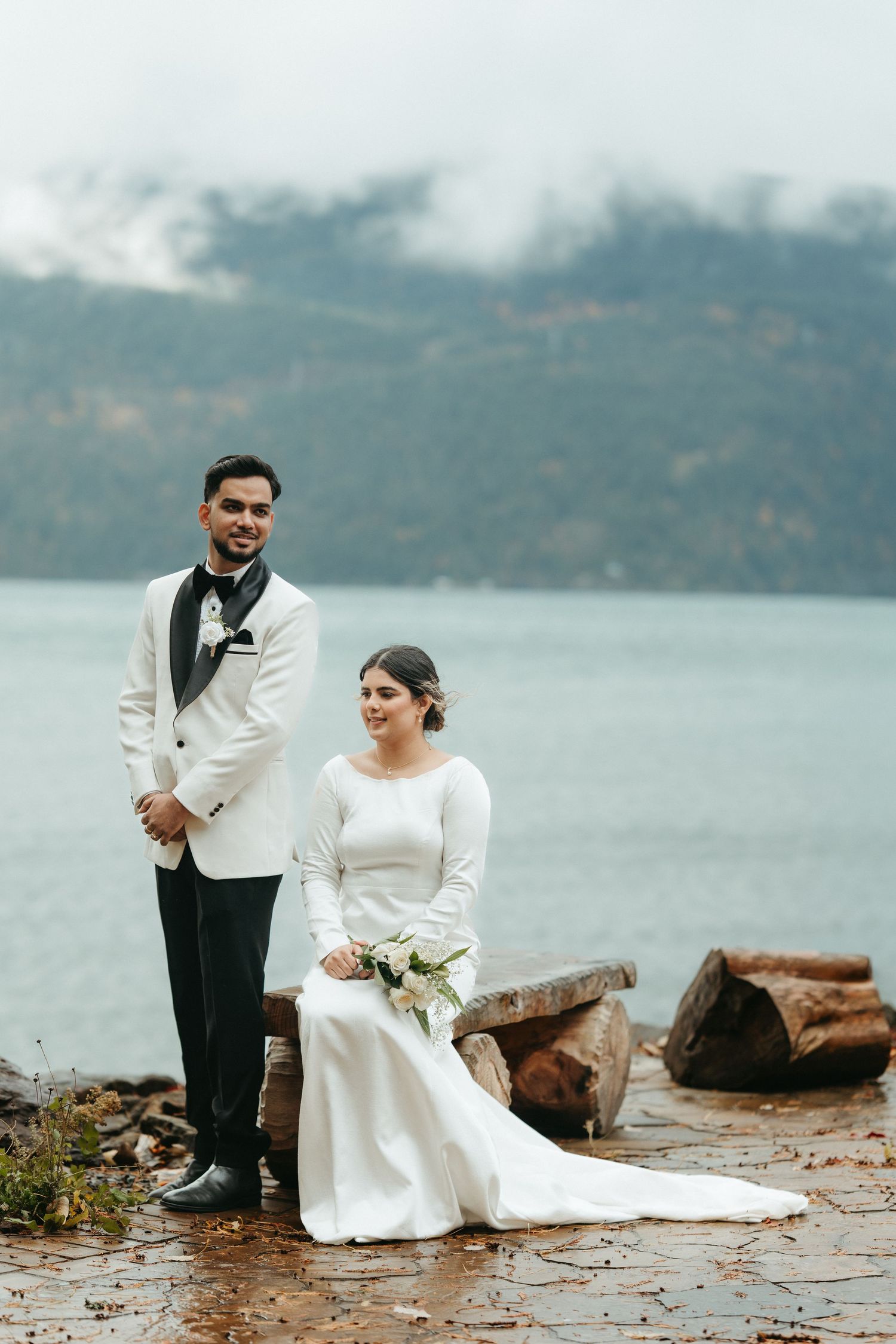A couple in wedding attire sits together on rocky shoreline with misty mountains and water in background.