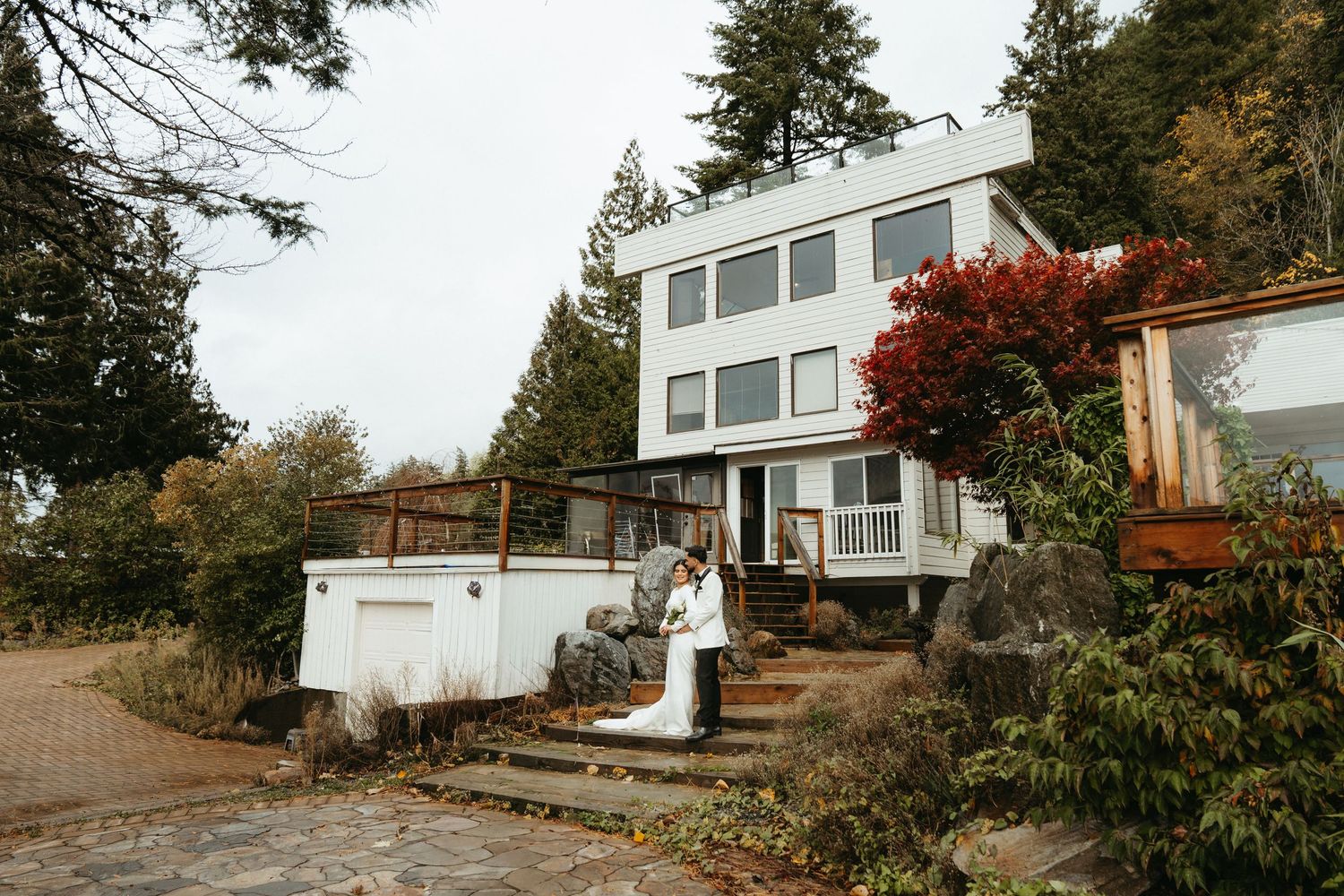 Modern white hillside home with large windows and natural landscaping surrounded by autumn foliage and trees.