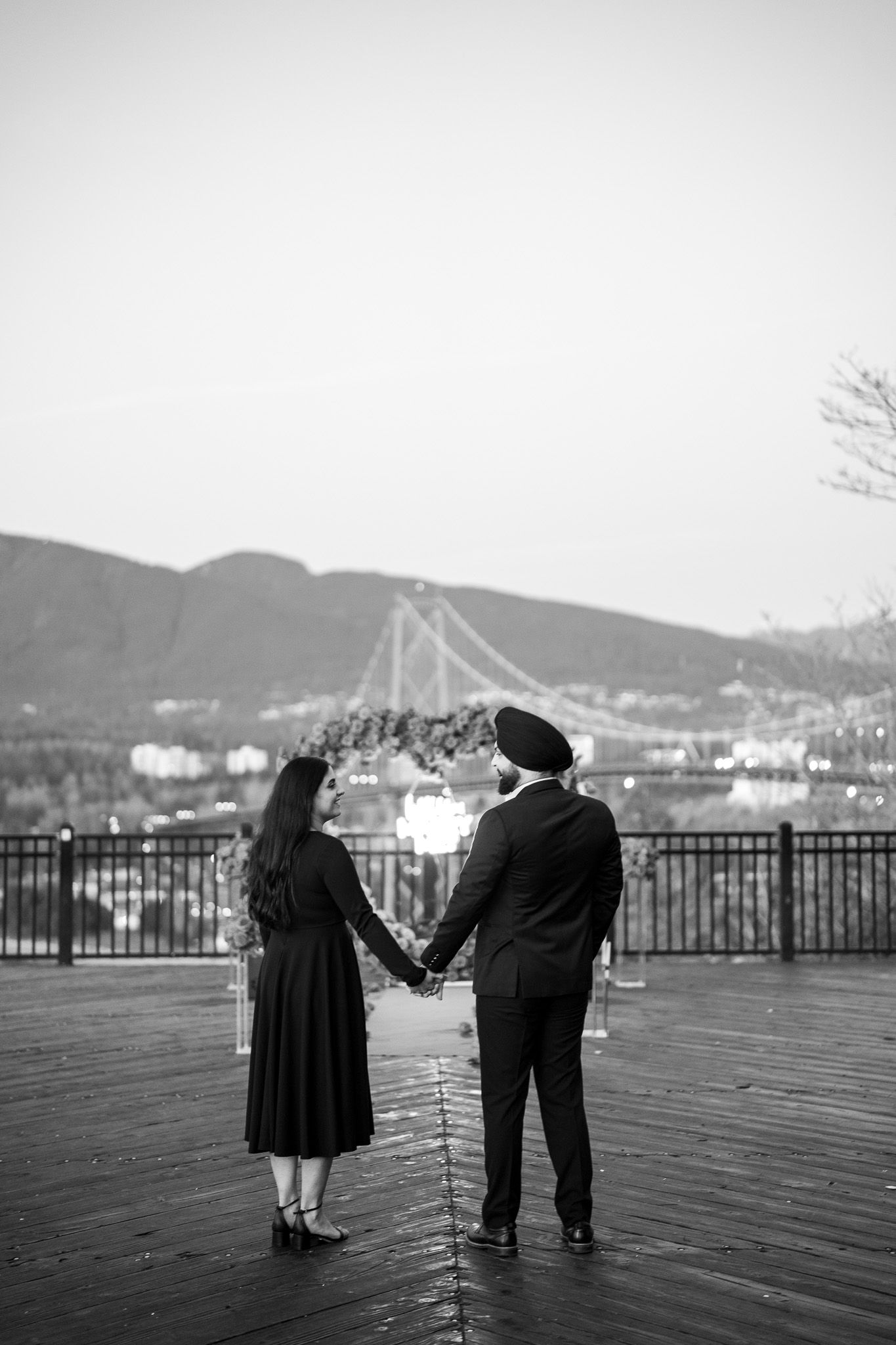 An elderly couple stands together on a wooden boardwalk overlooking a scenic mountain and harbor view in black and white.