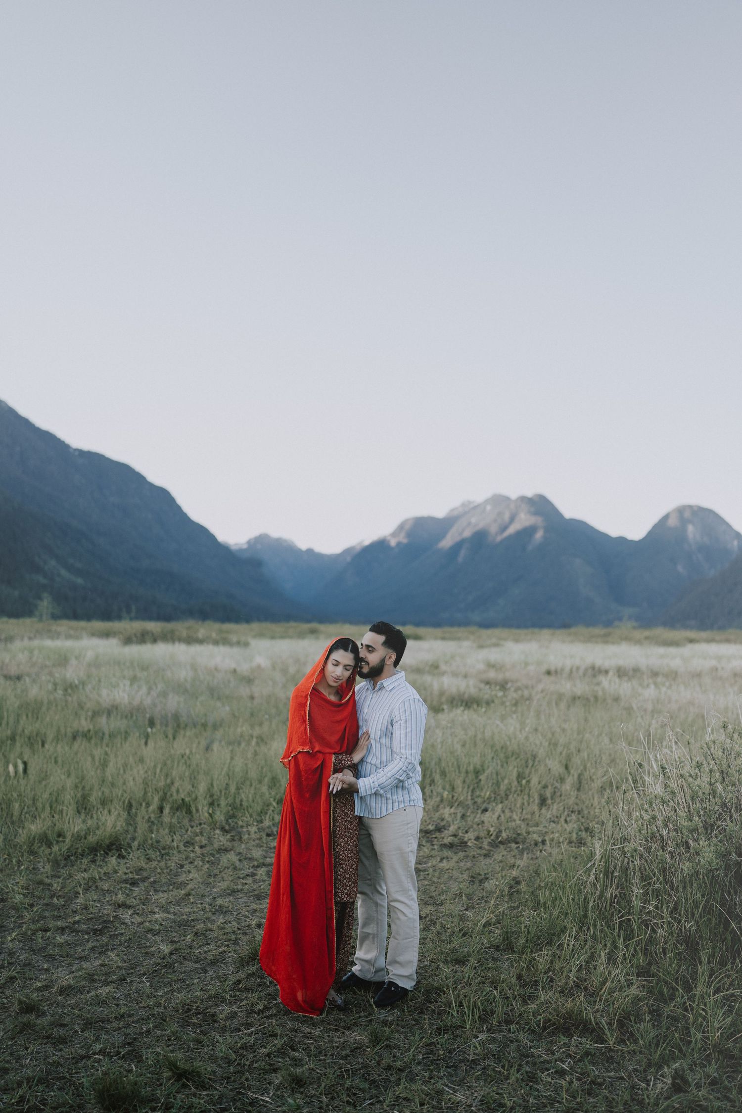 A couple stands together in a grassy meadow with majestic mountain peaks creating a dramatic backdrop in the distance.