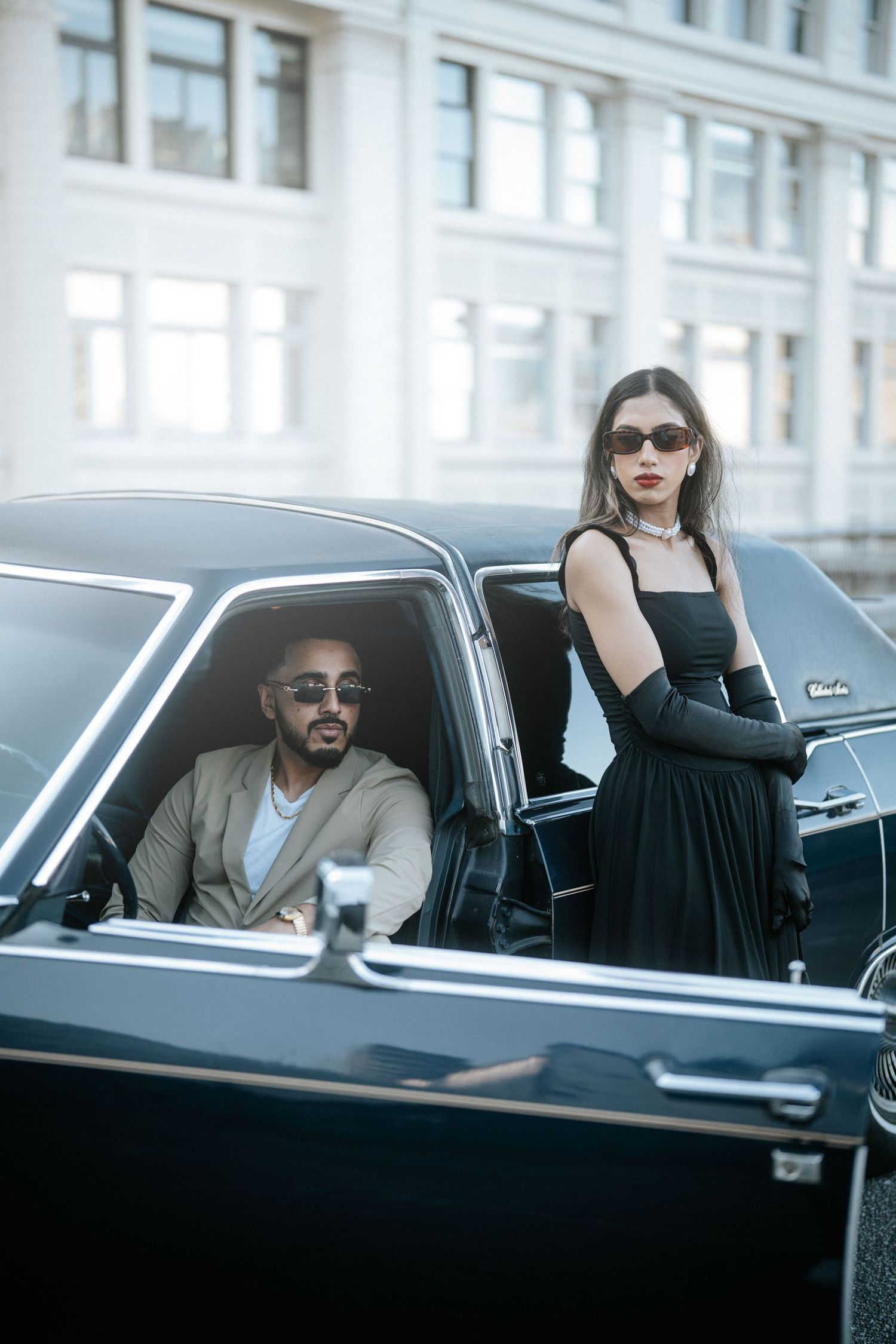 Stylish couple in black luxury car posing for fashion photoshoot against white building exterior.